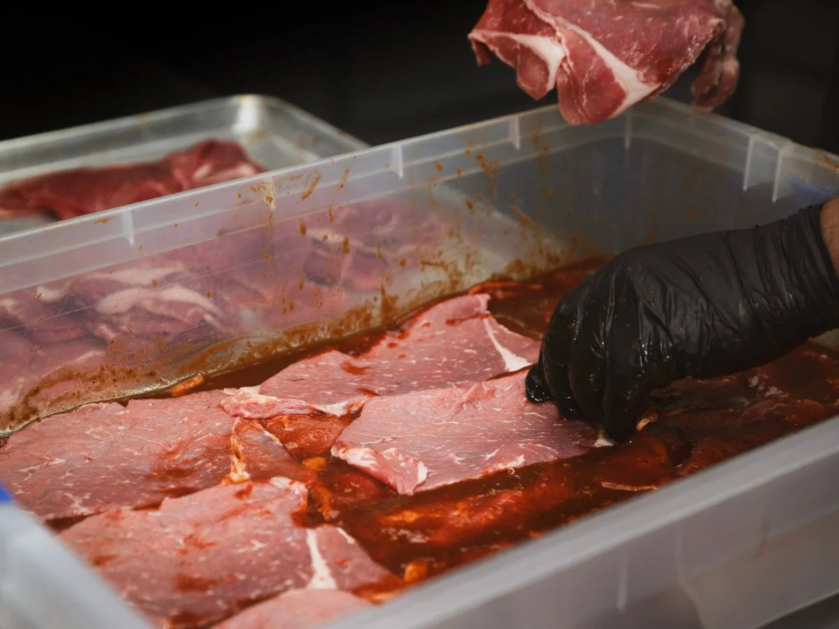 Someone wearing a black glove placing raw beef steak into a marinade in a plastic container.