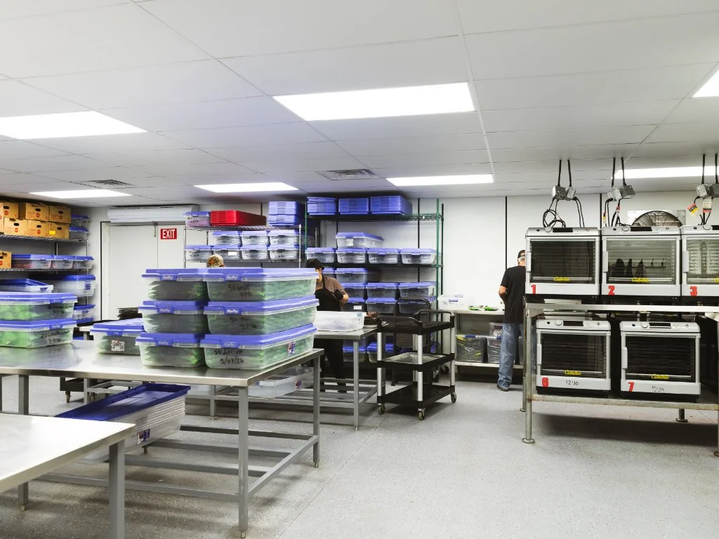A commercial kitchen or food storage room with stainless steel tables, shelves filled with food containers, and people working in the background.
