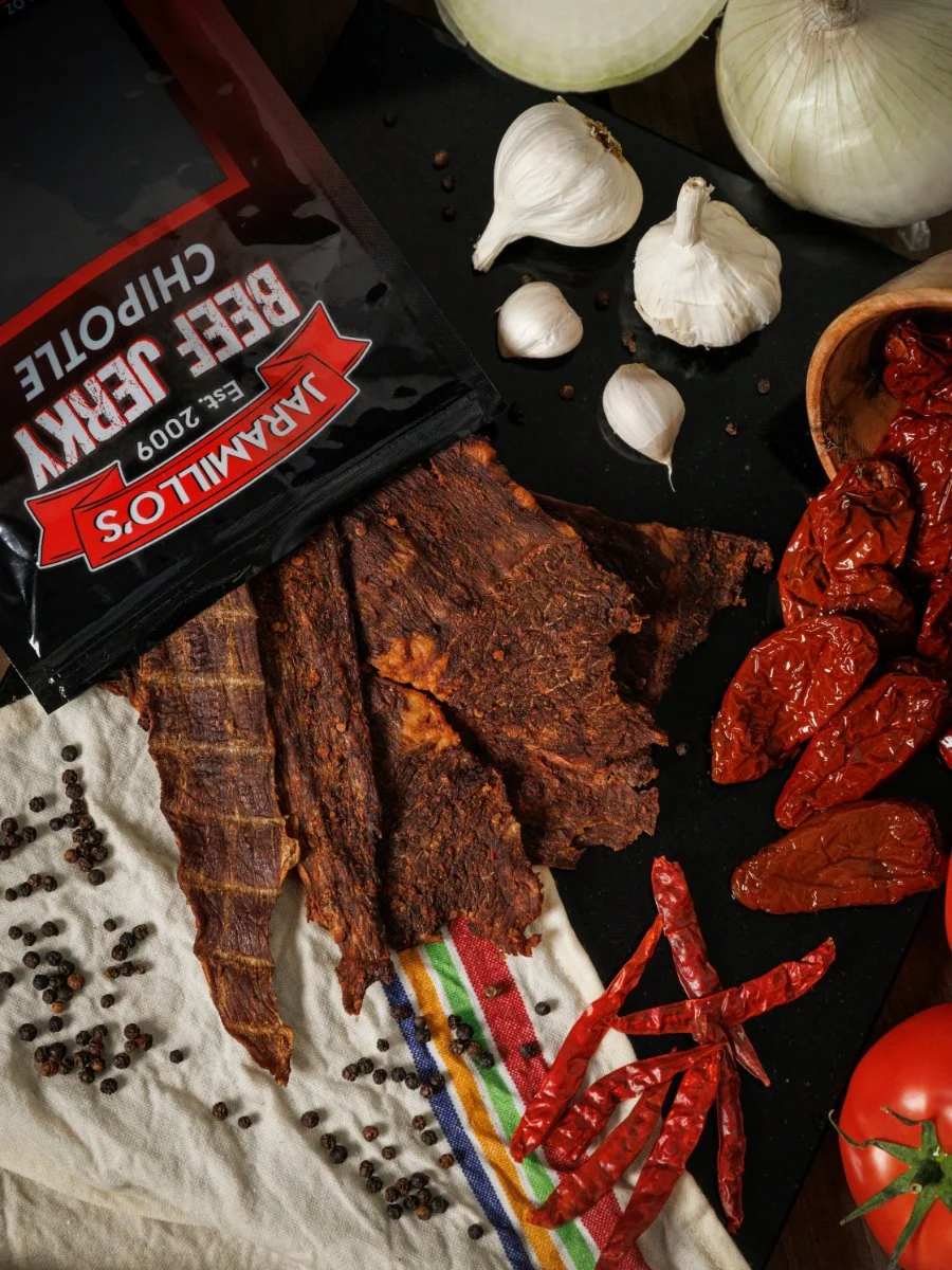 Dried beef jerky, garlic bulbs, dried red chili peppers, tomato, and a bag of Jack Daniel's beef jerky on a black surface with scattered black peppercorns on a white cloth.