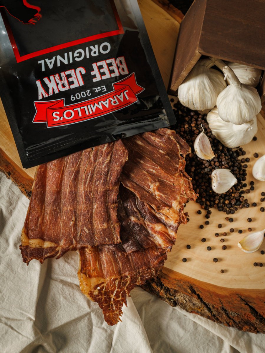 Dried beef jerky on a wooden cutting board, surrounded by garlic bulbs, garlic cloves, black peppercorns, and a packet of Jarmillo's Big Easy Jerky, Original flavor.