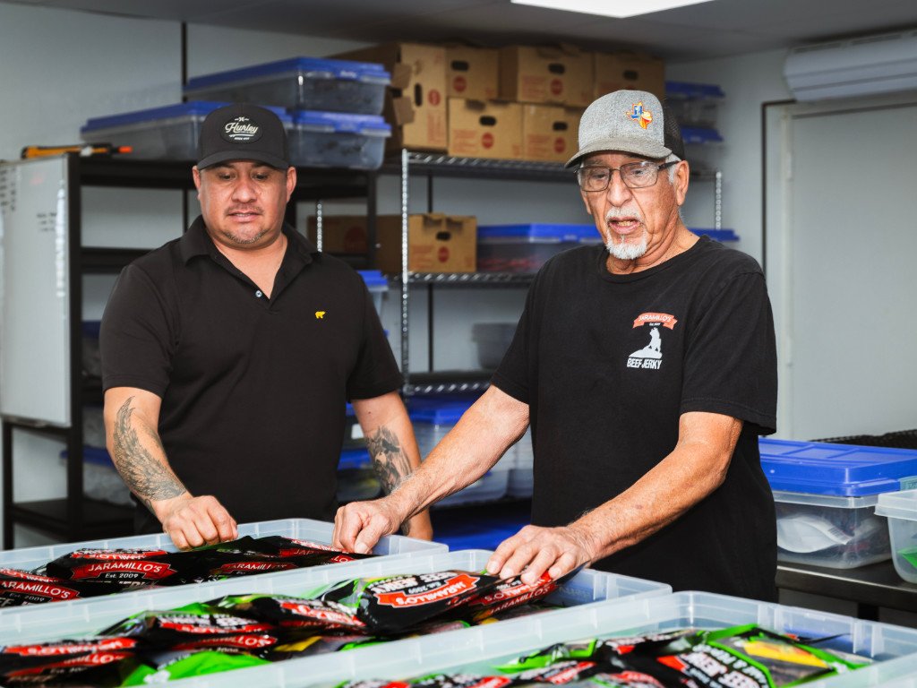 Two men inspecting packaged beef jerky in a storage room with shelves of boxes and containers.
