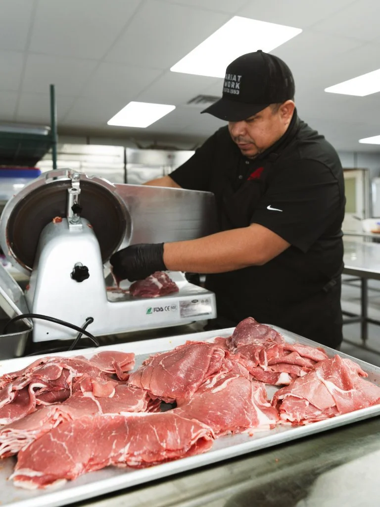 A man using a meat slicer to prepare cuts of raw meat in a professional kitchen.