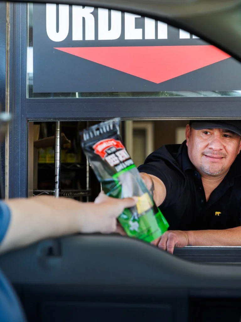 A fast food worker in a black shirt and hat is handing a bag of potato chips to a customer at a drive-thru window.