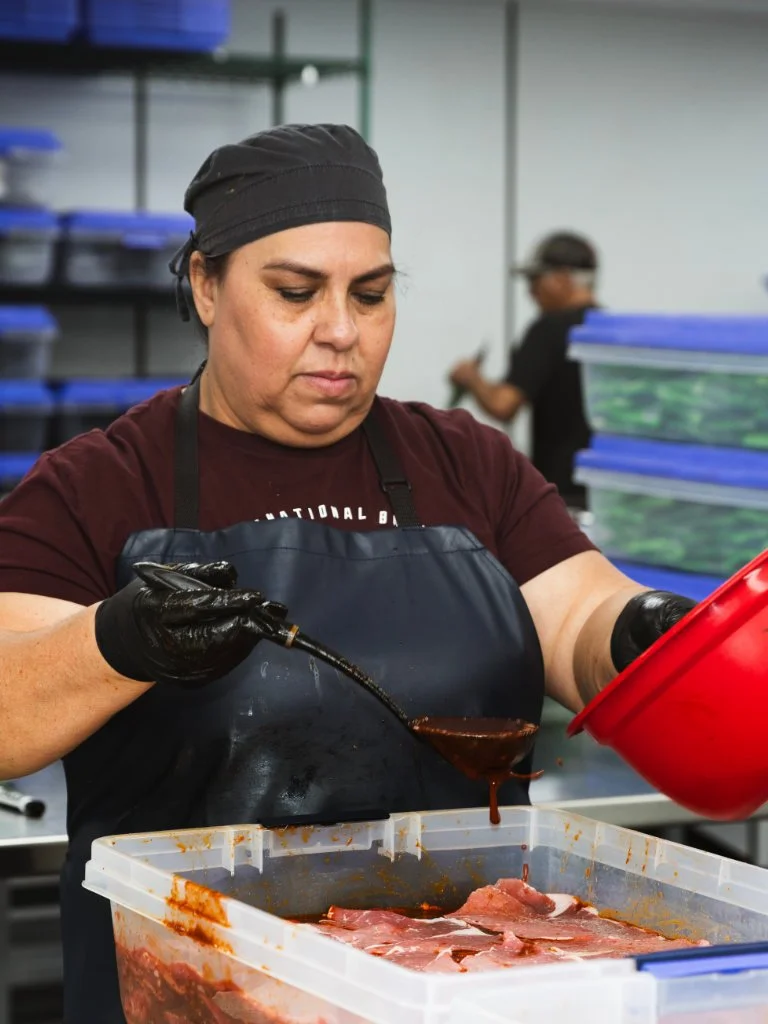 A woman wearing a black cap and apron, using a spoon to pour marinade over raw meat in a plastic container, with a background of blue storage bins in a commercial kitchen or food processing facility.