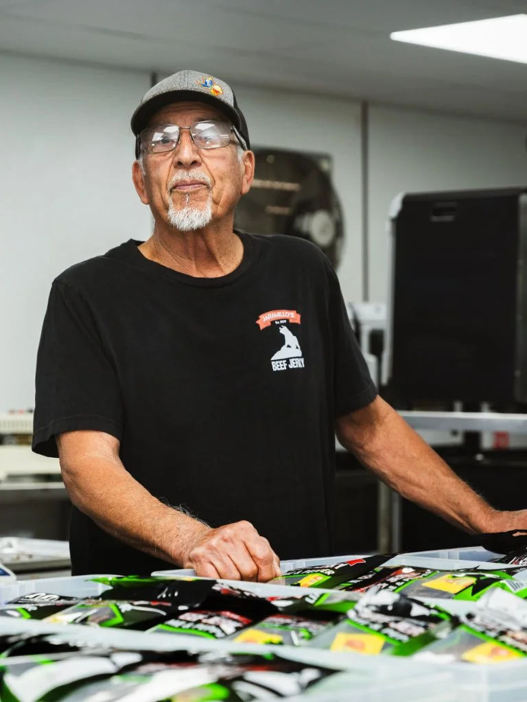 An elderly man with glasses, gray beard, and mustache, wearing a black T-shirt with a logo and a gray cap, stands behind a table with numerous snack bags in a room with a white wall and electronic equipment.