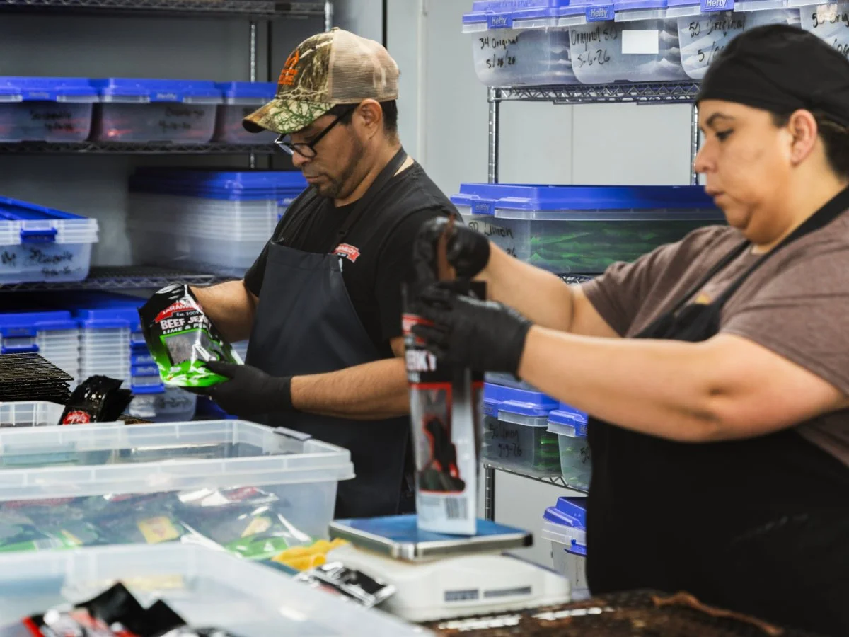 Two people working in a storage room or warehouse, packing or organizing items. The man on the left is wearing a camouflage cap, glasses, and a black apron, holding a packaging bag. The woman on the right is wearing a black cap, black gloves, and also handling packaging. Shelves behind and in front contain plastic storage bins with labels.