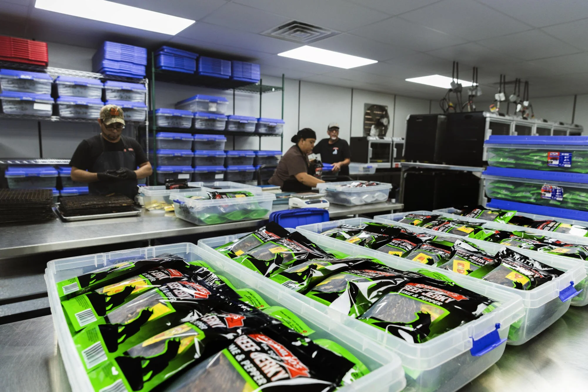 Workers in a supply room organizing black and green packaging in clear plastic bins, with shelves holding blue and red containers in the background.