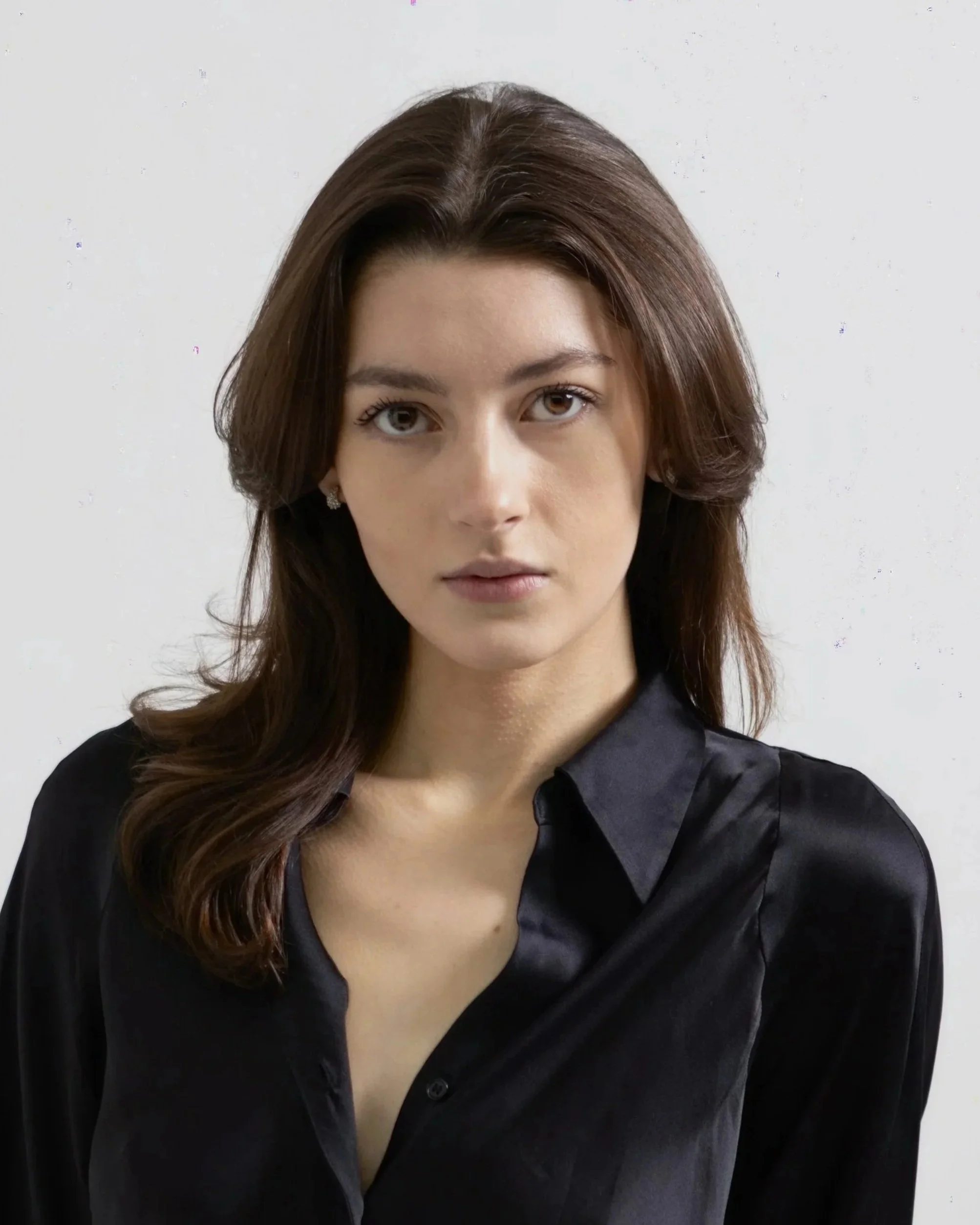 A woman with shoulder-length brown hair wearing a black satin blouse and earrings, standing against a plain white background.