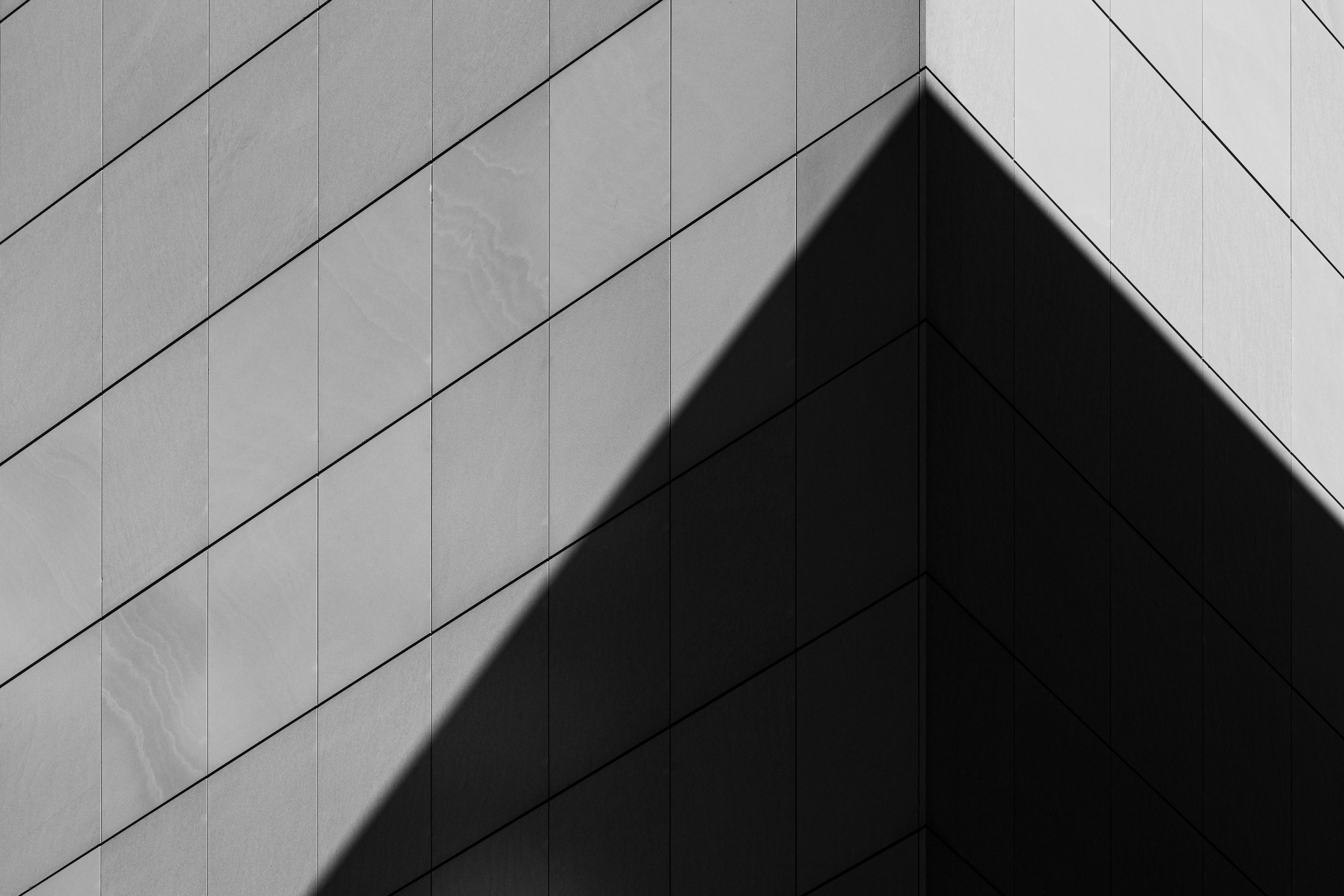 Vista en ángulo de la esquina de un edificio moderno con paredes de baldosas grises y sombra angular proyectada sobre ellas.