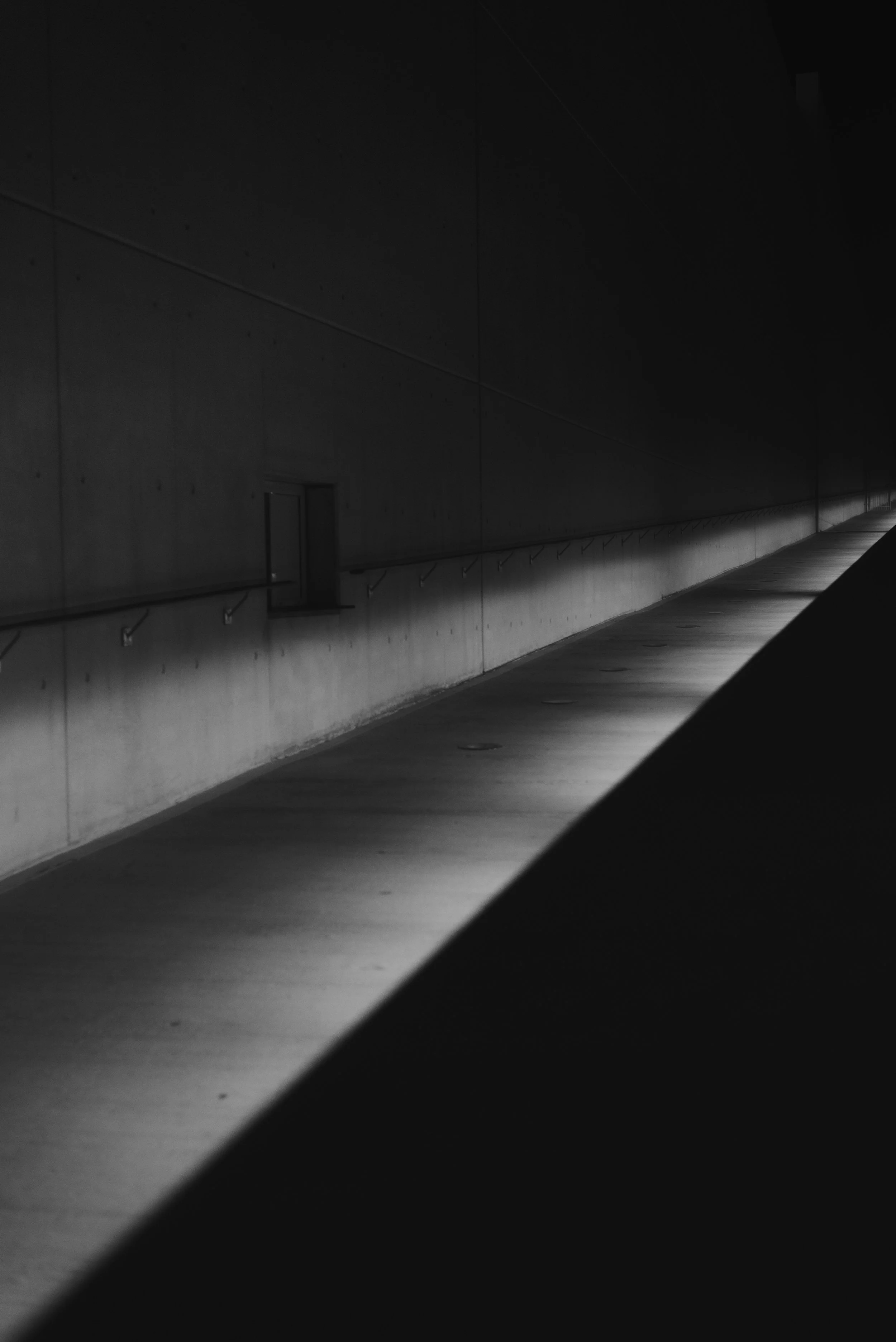 Pasillo oscuro con luz en una de sus paredes, en tonos grises y negros.