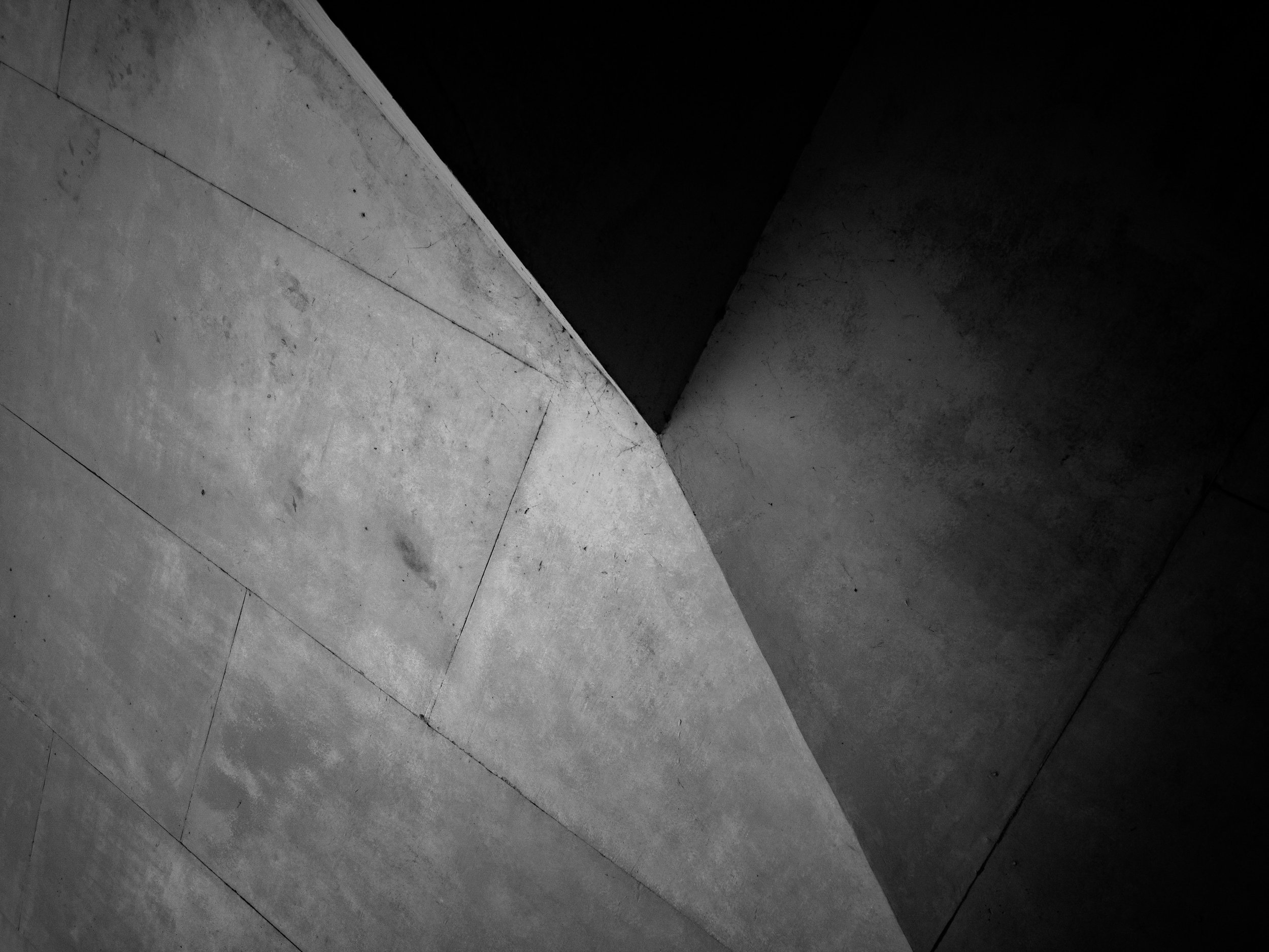 Una imagen en blanco y negro de una esquina de un espacio interior con paredes de concreto, con líneas diagonales y diferencias de iluminación que crean sombras y contrastes.