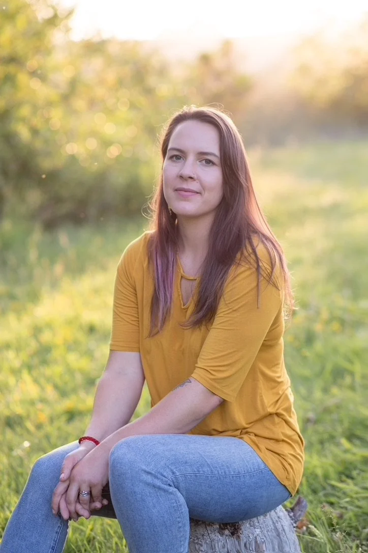 A young woman with long brown hair and purple tips, wearing a mustard yellow t-shirt and light blue jeans, sitting on a tree stump outdoors in a grassy area with trees in the background during what appears to be late afternoon or early evening.