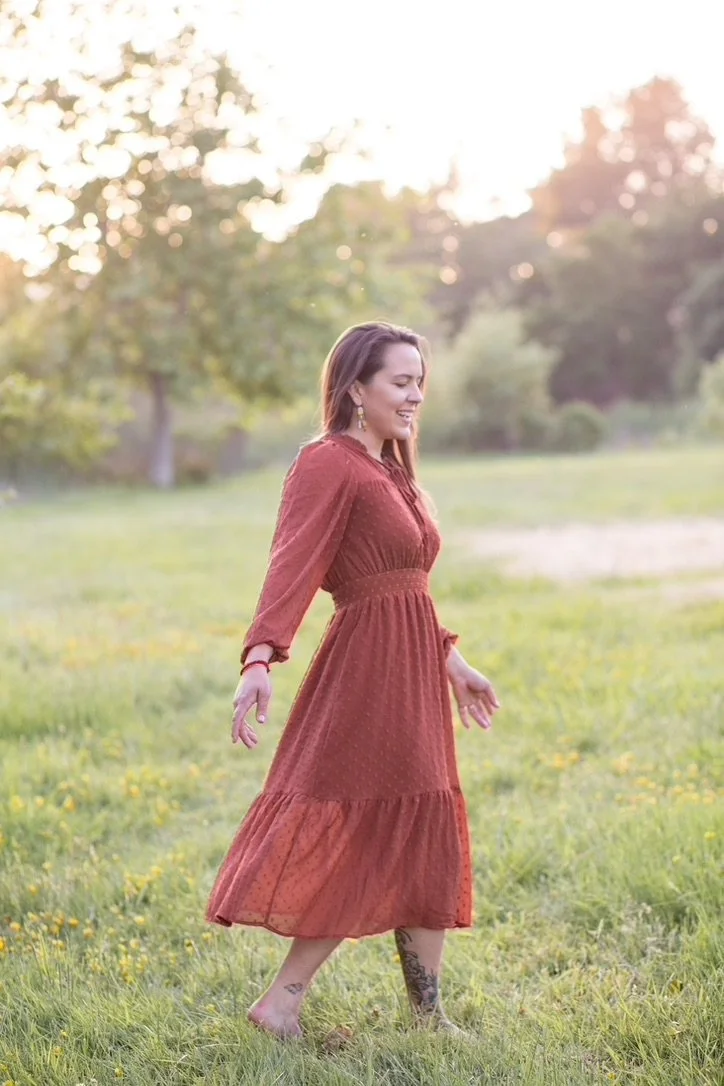 A woman with long brown hair, wearing a rust-colored, long-sleeved dress, is walking barefoot on green grass in a field with yellow flowers, during sunset or late afternoon, with trees in the background.