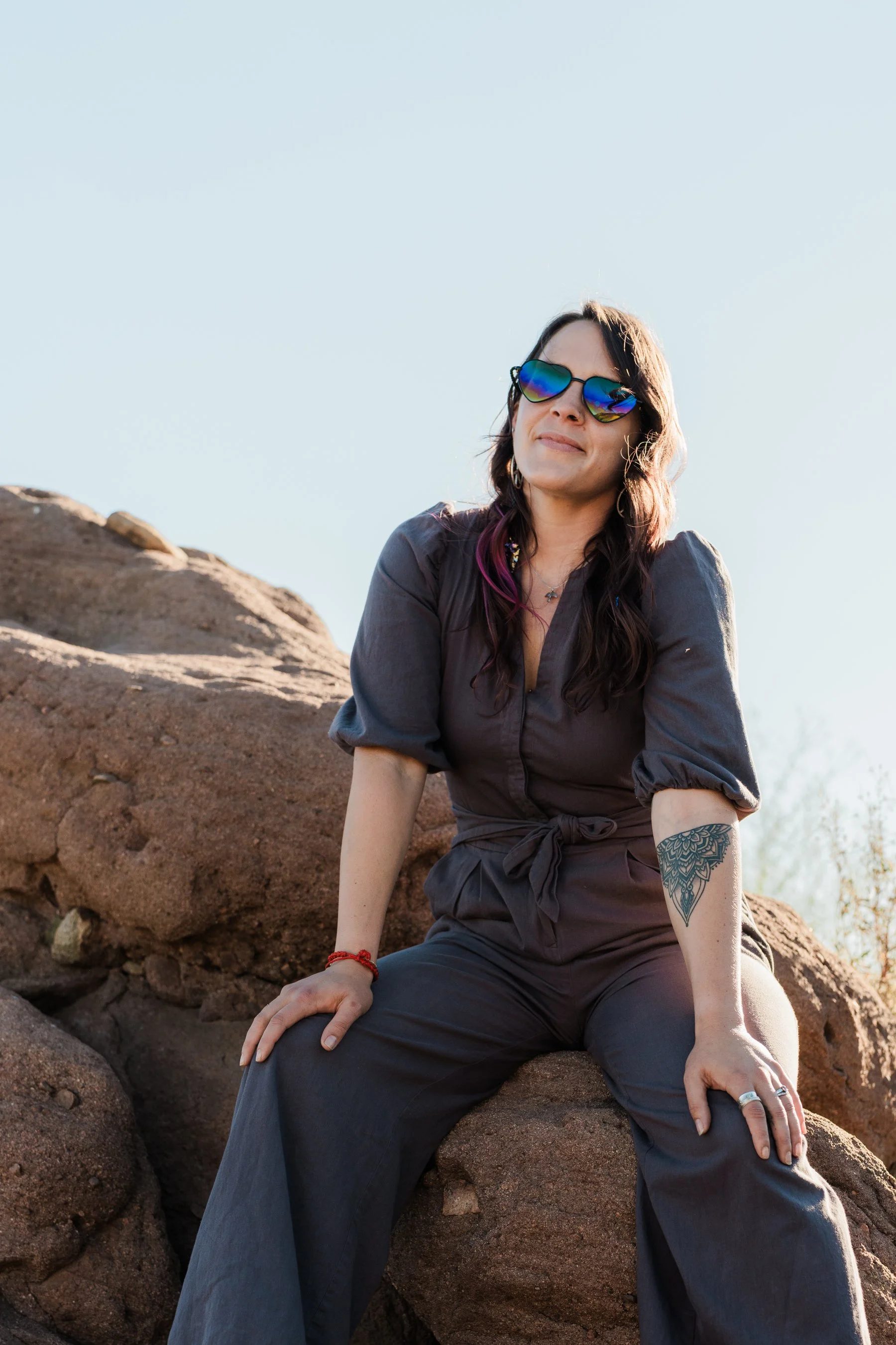 A woman sitting on rocks outdoors, wearing sunglasses, a gray jumpsuit, and displaying a tattoo on her right forearm.