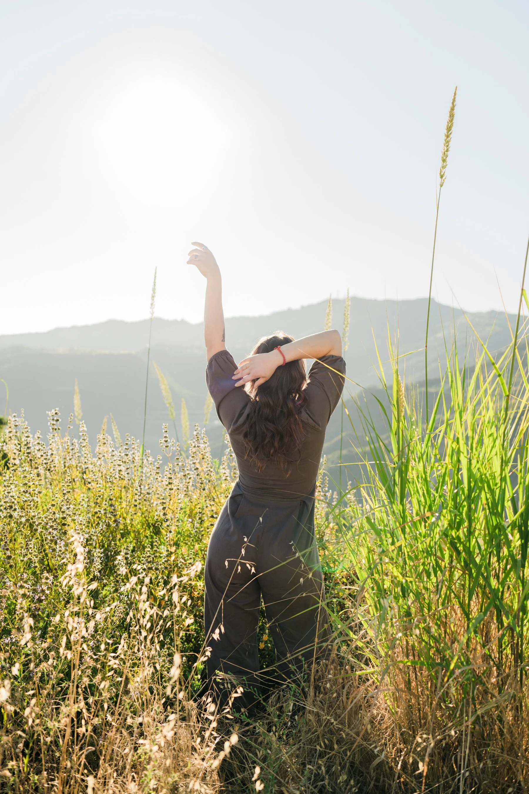 Woman standing in a grassy field with her back to the camera, raising one arm and with her other arm touching her head, during bright daylight with hills in the background.