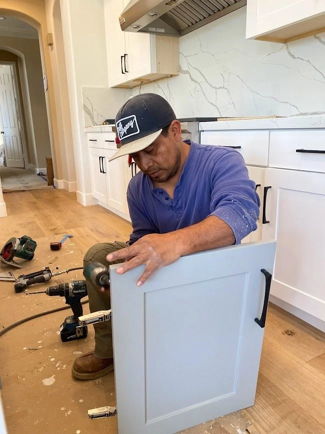 A man kneels on the floor, working on a white kitchen cabinet with a power drill, surrounded by scattered tools.
