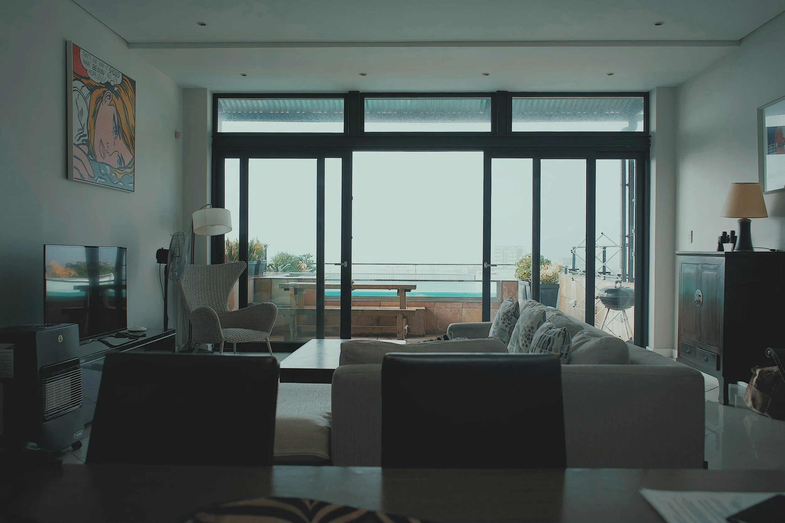 Living room with sliding glass doors leading to balcony, view of sky and distant buildings, furniture including a sofa, armchair, and TV, with decorative elements like a painting and a lamp.