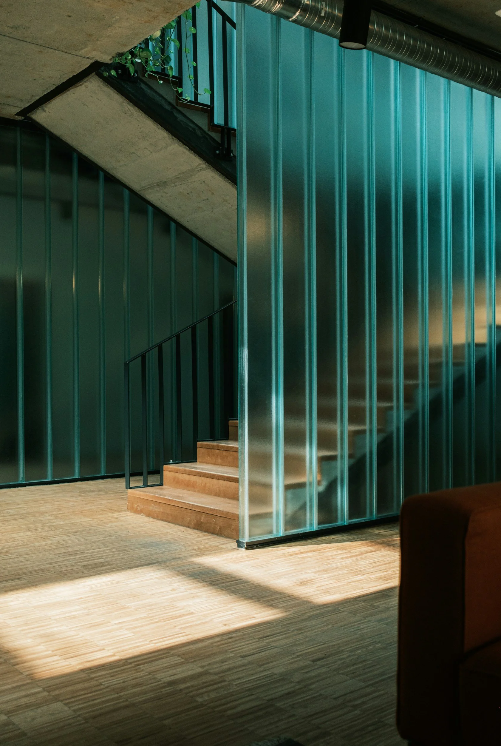 Interior view of a modern staircase with wooden steps, black handrails, green vertical metal wall panels, a concrete ceiling with exposed ductwork, and sunlight casting shadows on wooden flooring.
