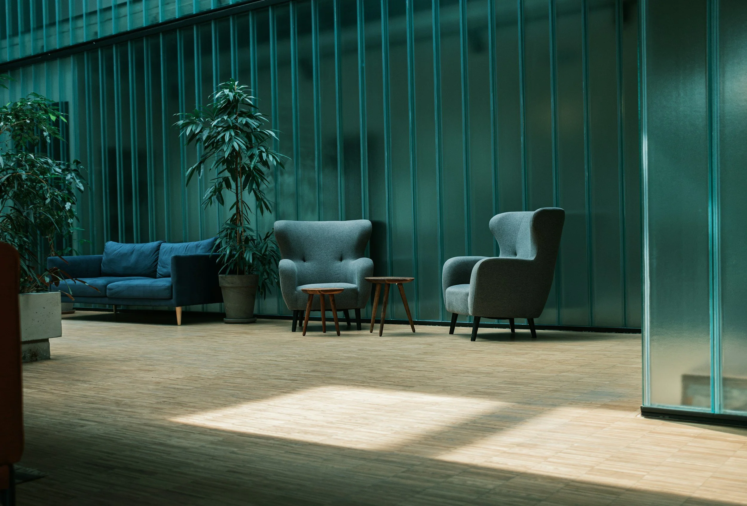 Modern lounge with blue sofa, two gray high-back armchairs, two small wooden side tables, potted plants, and a teal-colored wall with vertical panels.