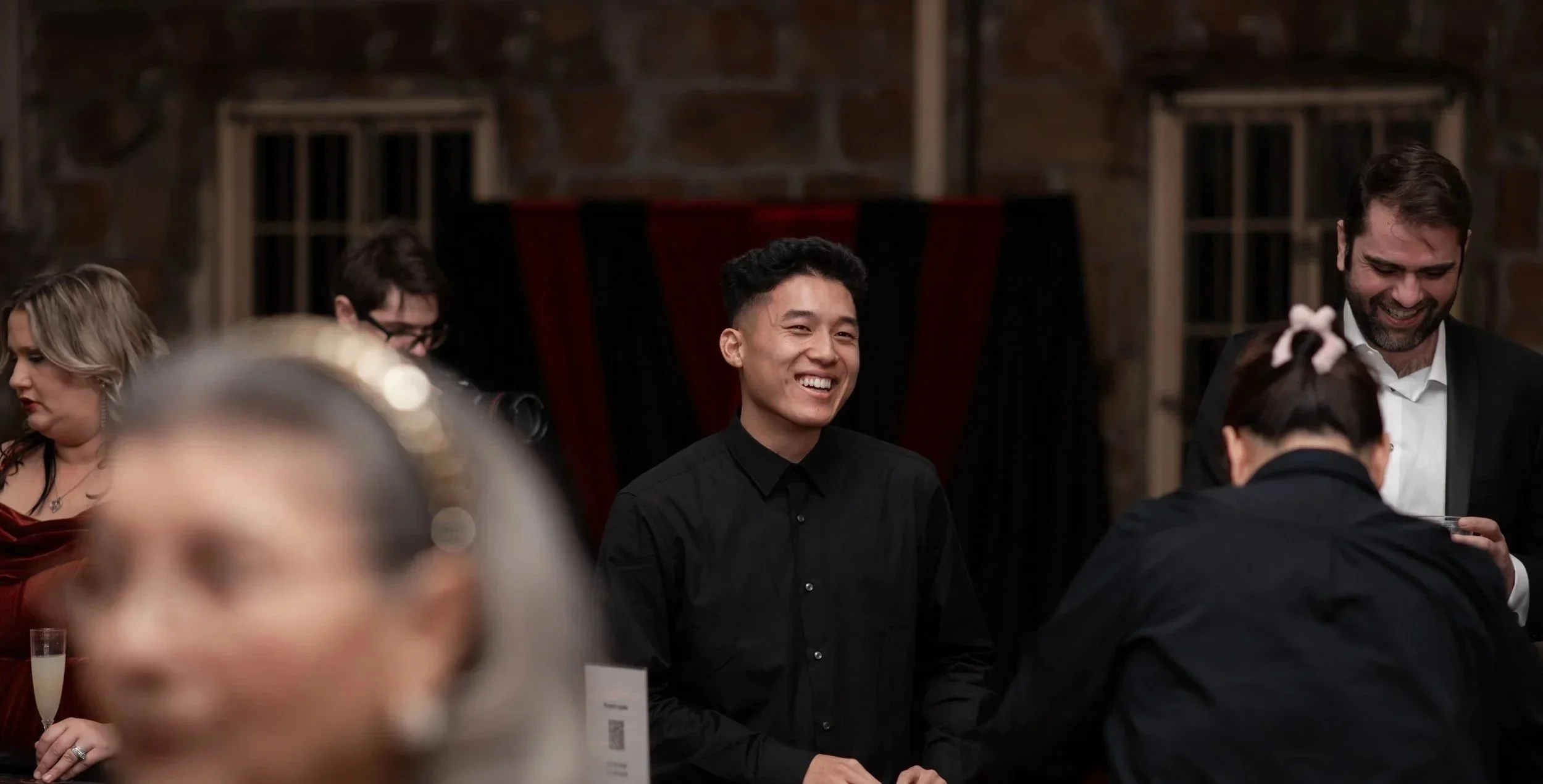 A group of people at a social gathering, with a smiling man at the center, wearing a black shirt, in a warmly lit room with brick walls.