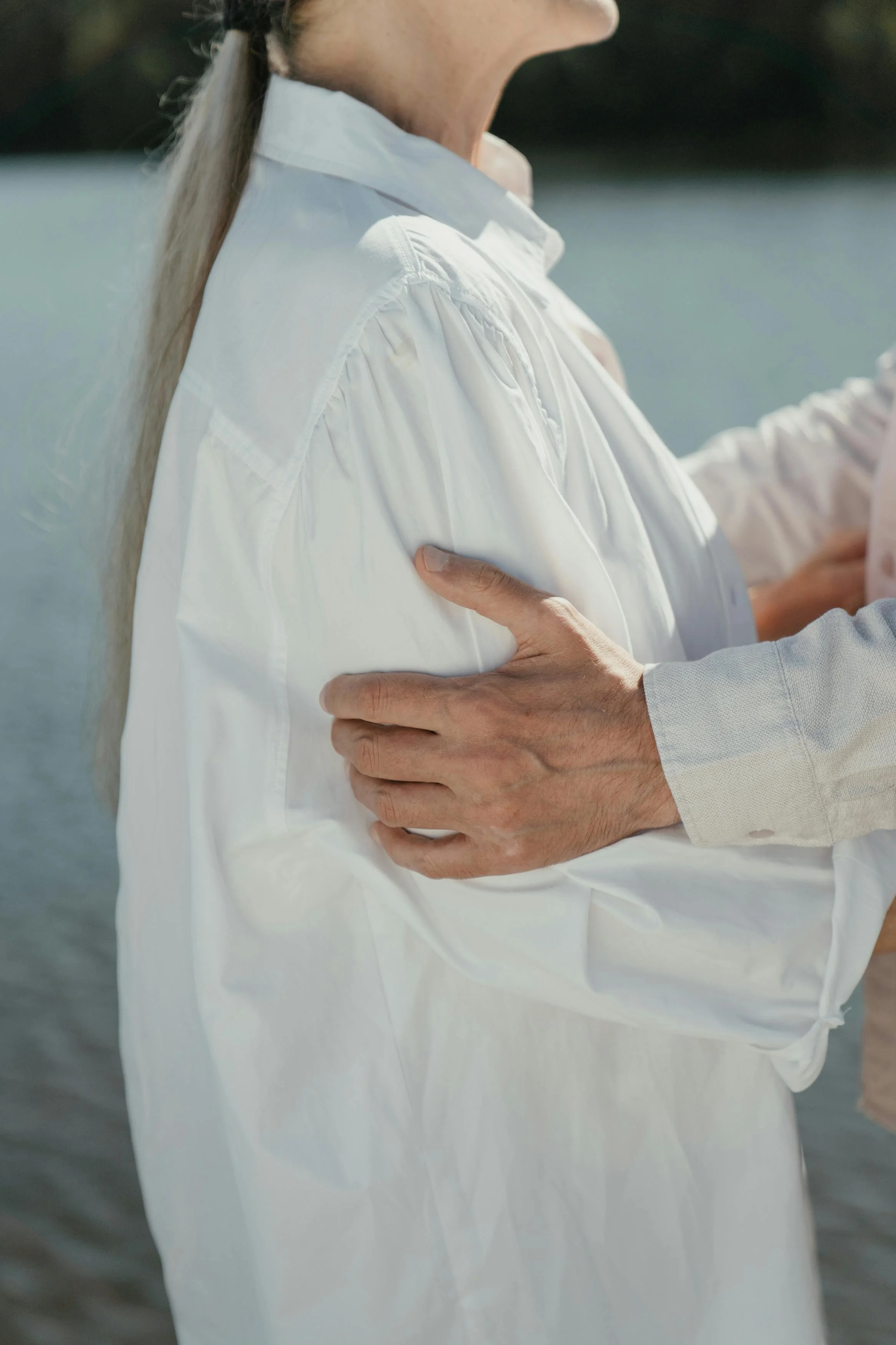 Close-up of a person in a white shirt with long blonde hair beside a body of water, with another person touching their arm.