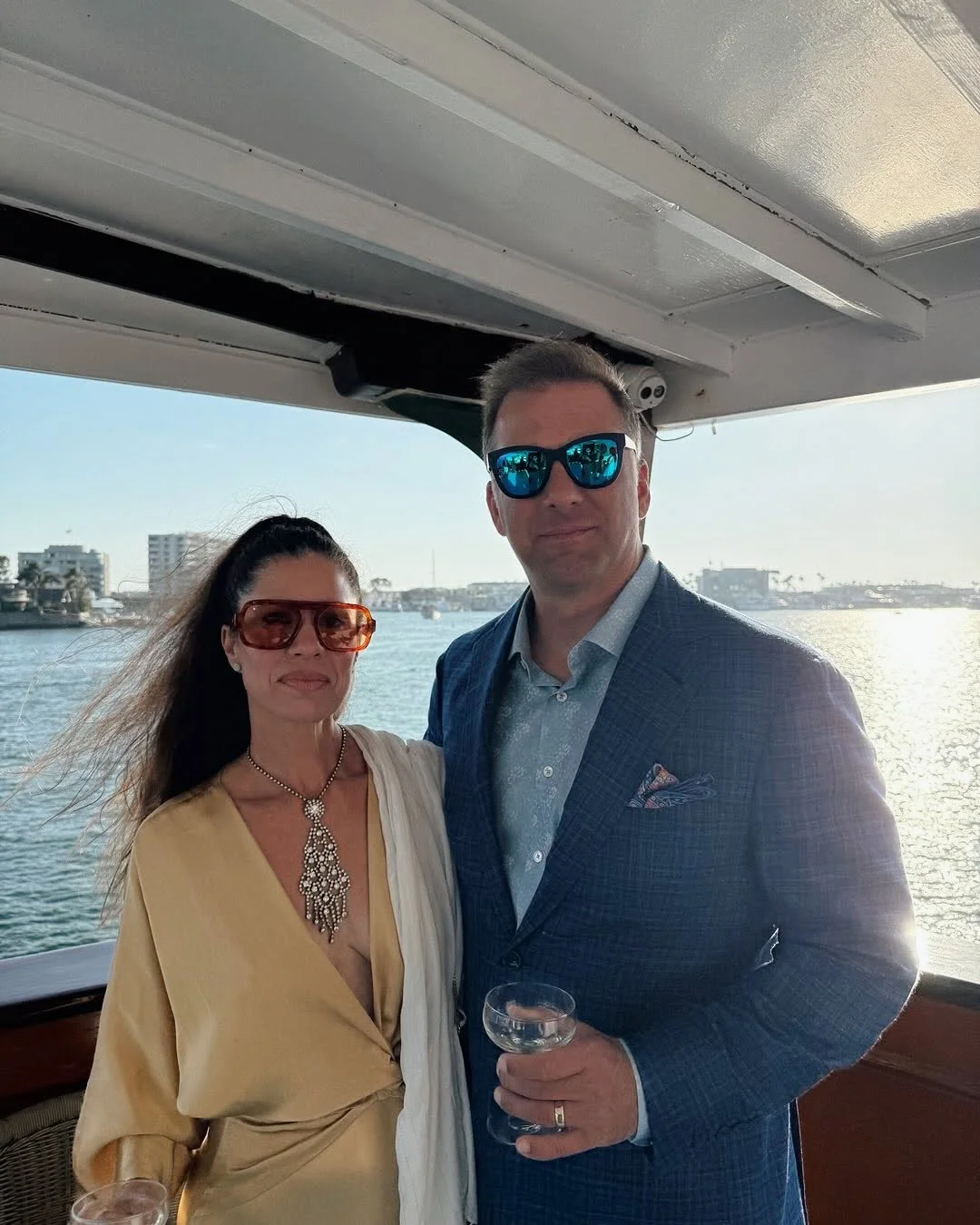 A man and woman on a boat, dressed upscale, wearing sunglasses, with a body of water and buildings in the background, during sunset.