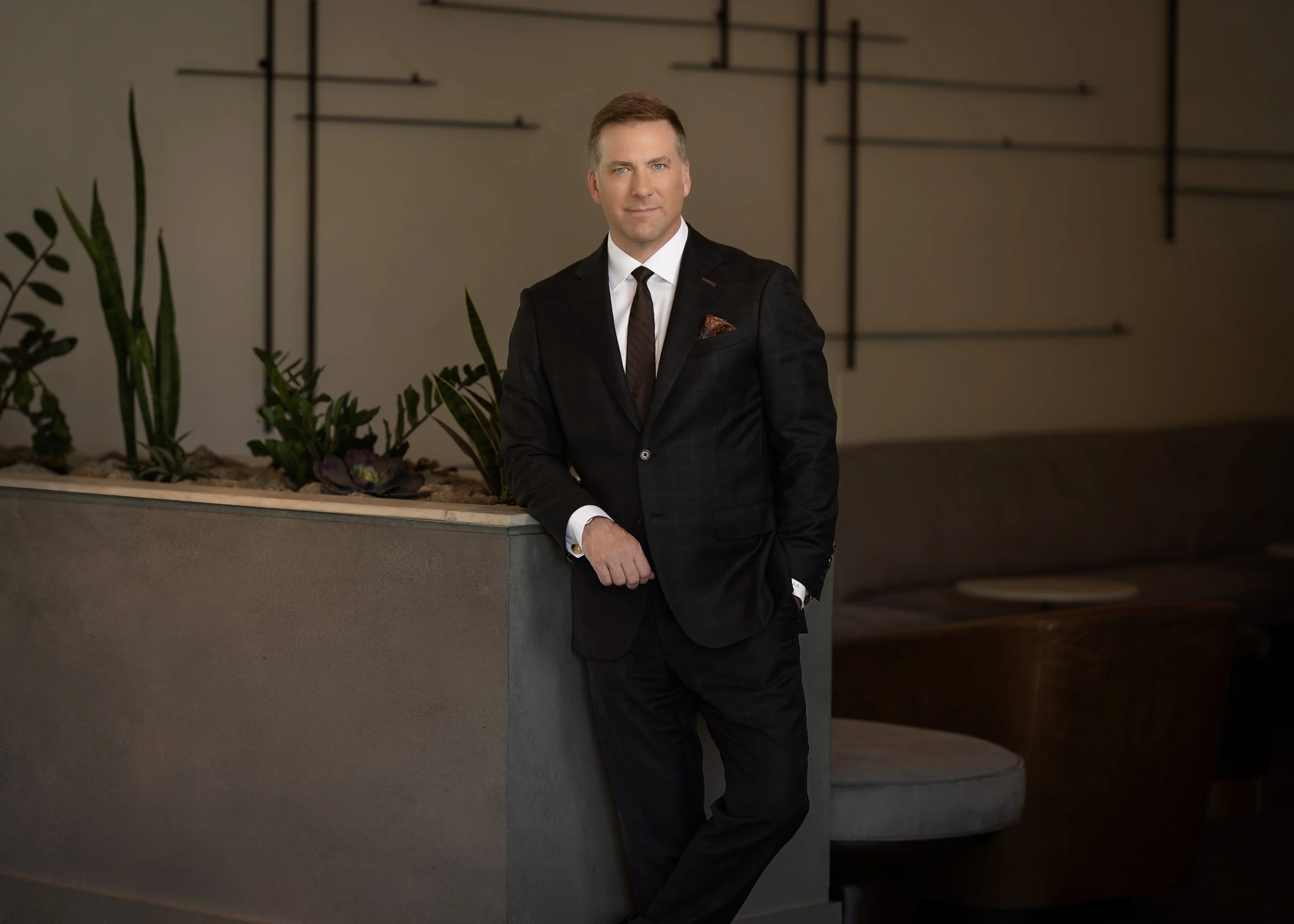 A man in a black suit, white shirt, and black tie standing in an interior space with modern decor, including plants and wall art.