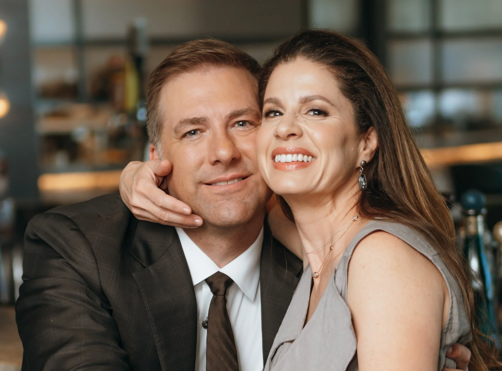 A man in a suit and a woman with long hair smiling and hugging each other in an indoor setting.