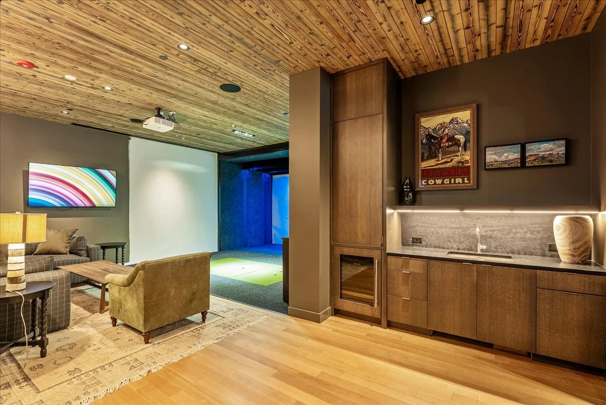 Living room with wooden ceiling, modern furniture, wall-mounted TV displaying colorful rainbow pattern, and a dry golf putting green area. A kitchenette with wooden cabinets, a stone backsplash, and decorative art including a vintage Utah Cowgirl pos