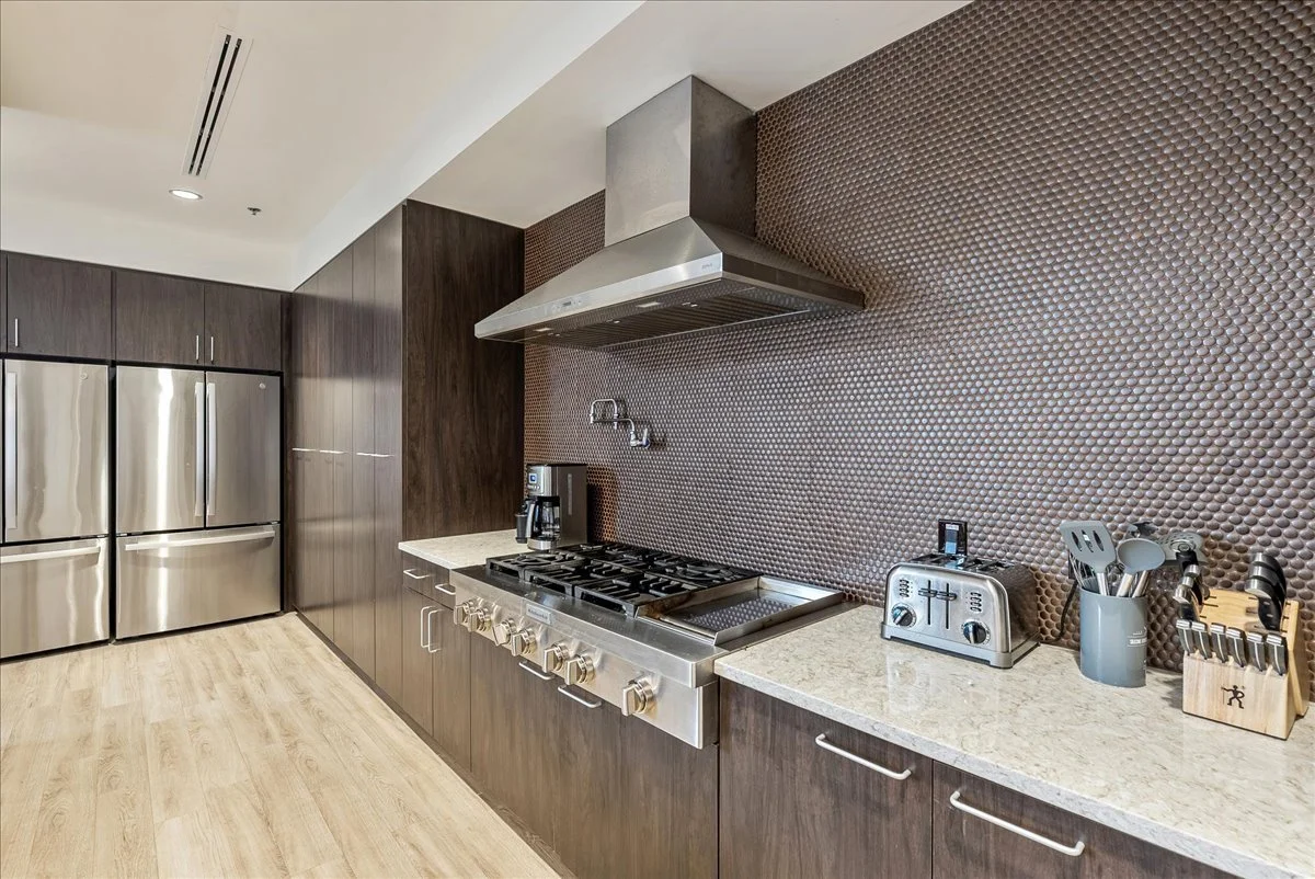 Commercial kitchen with dark wood cabinets, stainless steel refrigerator, gas stove with range hood, coffee maker, toaster, utensils, and cutting tools on countertop with textured brown backsplash.