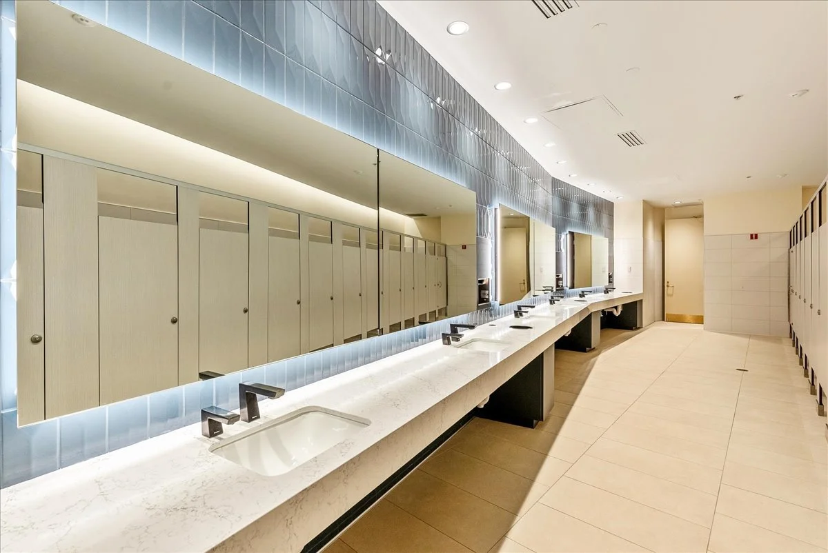 A public restroom with a long marble countertop, multiple sinks, and black faucets, mirrored cabinets above, and lockers to the side.