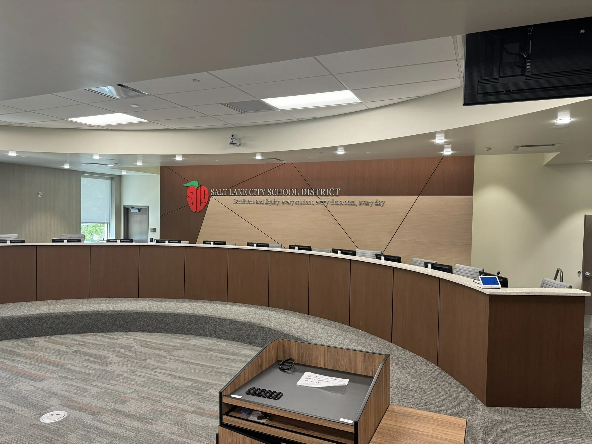 Interior view of a modern office or conference room with a curved wooden reception desk, a small table with a keyboard and papers, and a wooden wall with the Salt Lake City School District logo and slogan. Overhead lighting and a window are visible.