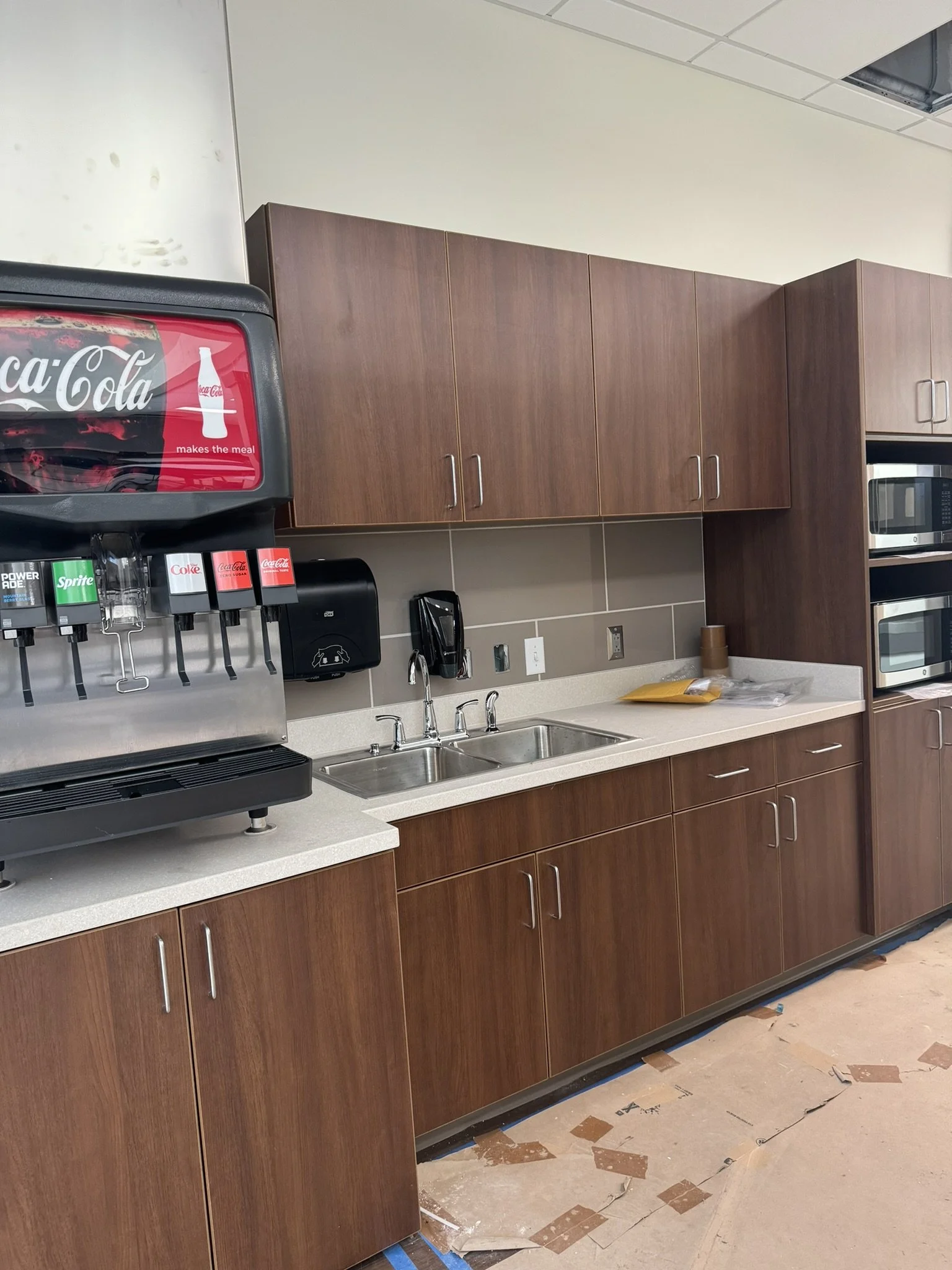 Cleaning or renovation in progress at a kitchenette area with wooden cabinets, a stainless steel sink, a beverage dispenser, and nearby oven and microwave. The floor shows signs of ongoing work with protective paper and tape.