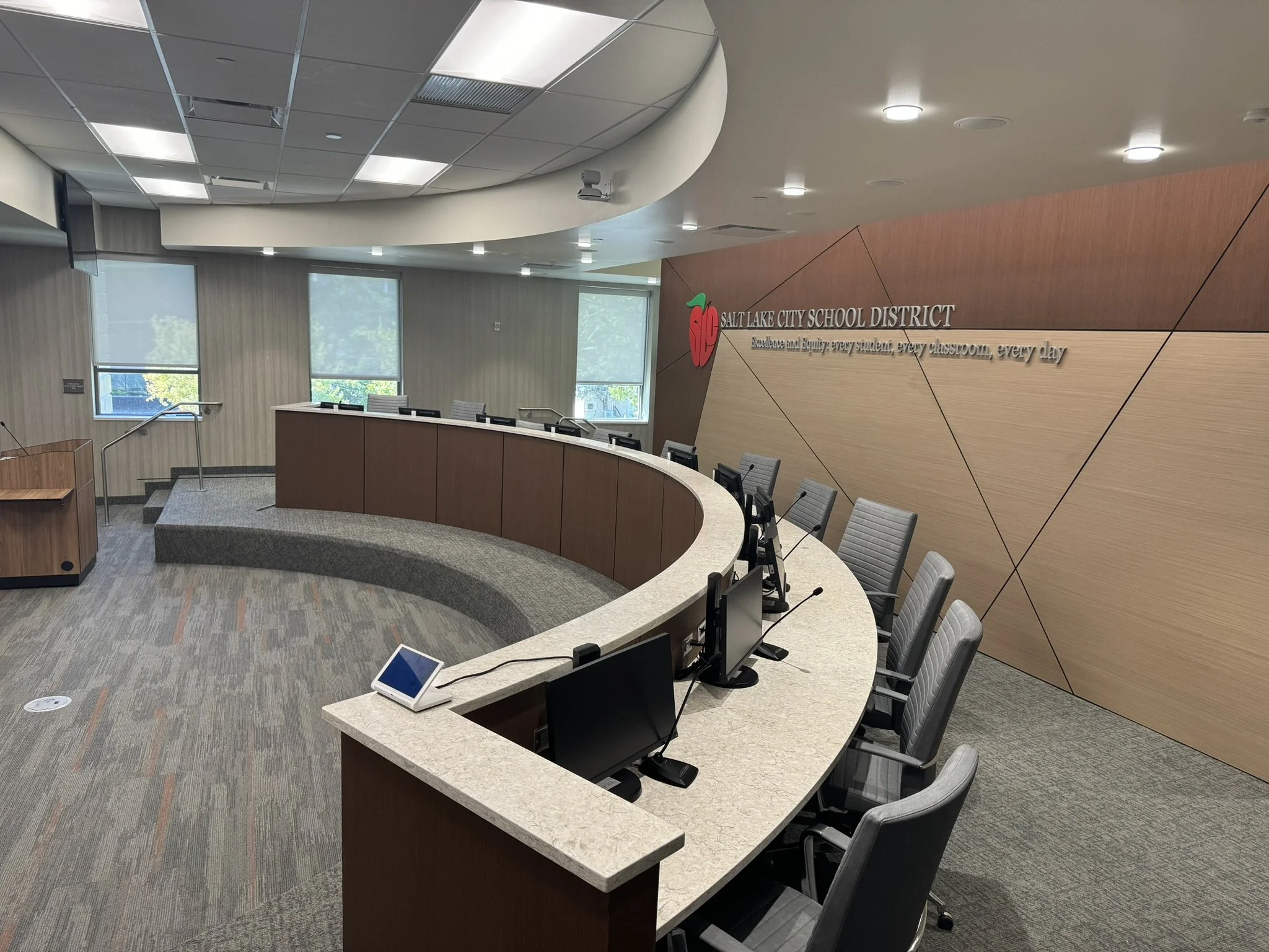 A modern conference room with curved wooden tables, ergonomic office chairs, and digital tablets on the tables. The room features large windows with blinds, a wood paneled wall with the Salt Lake City School District logo and motto, and a high ceilin