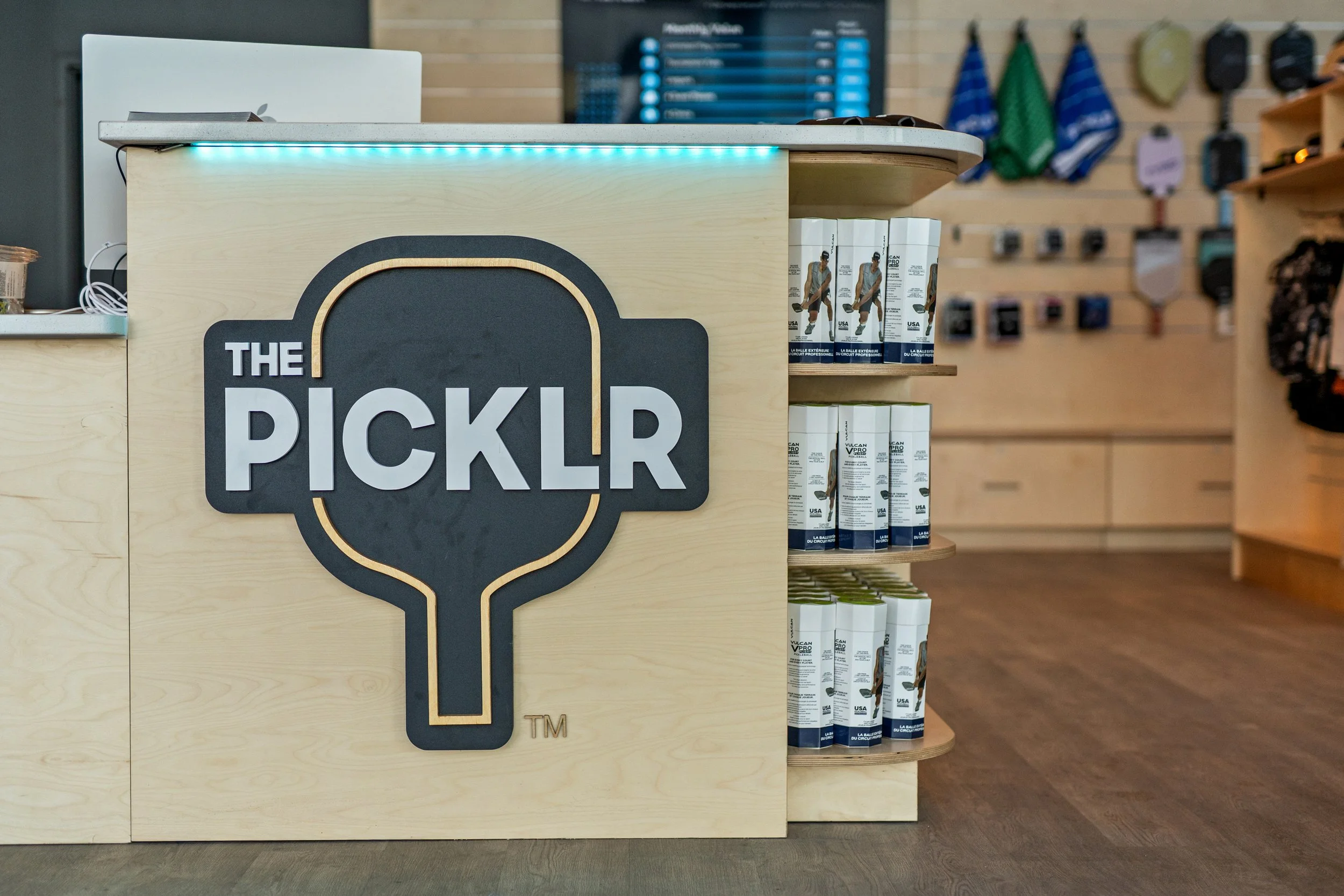 A retail checkout counter with a sign that reads 'The PickLR', positioned next to shelves display cans with a basketball player on the label. In the background, there are wall-mounted hooks displaying various bags and accessories, and a digital screen showing a purchasing or checkout menu.