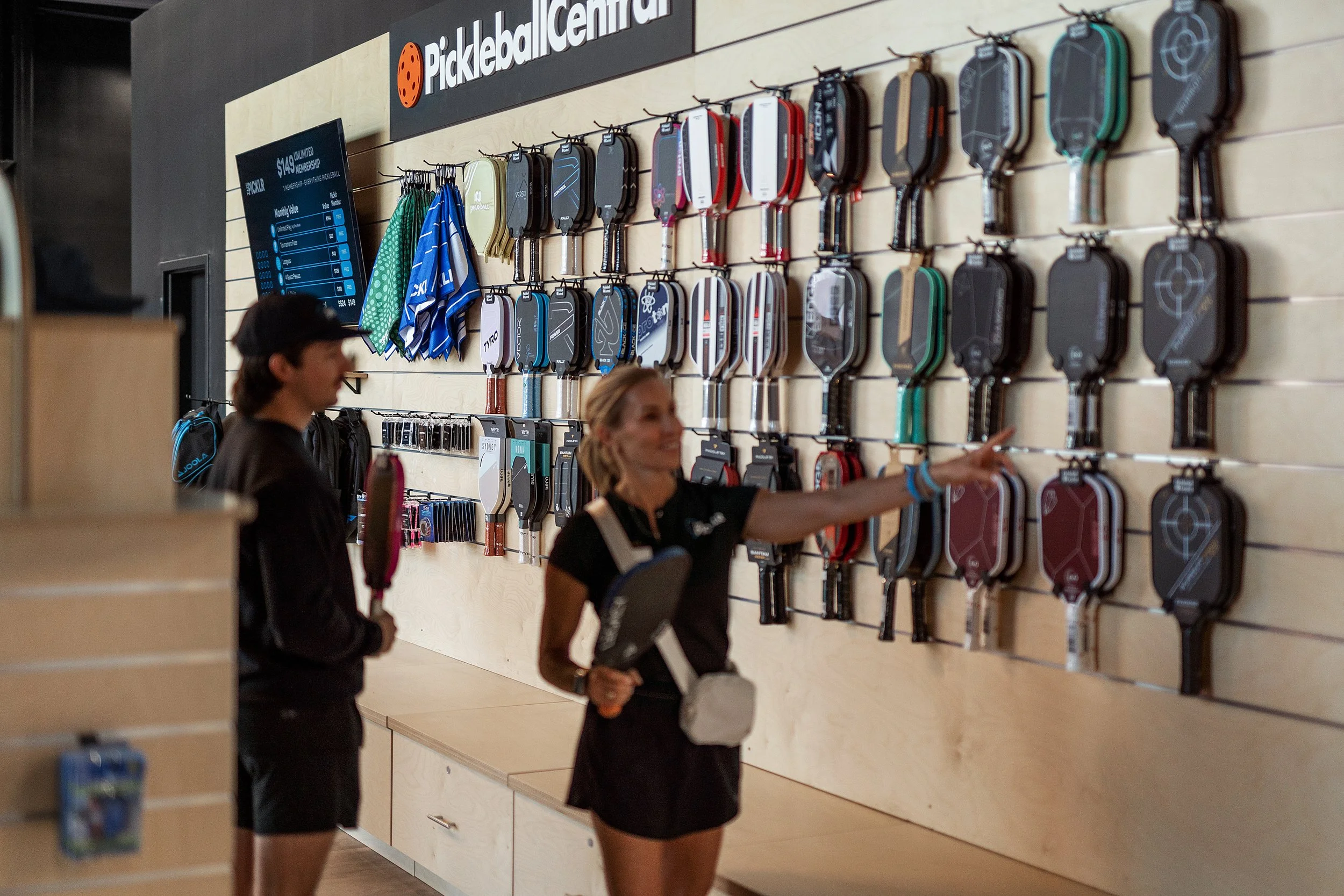 Two women shopping at a pickleball store with paddles displayed on the wall.