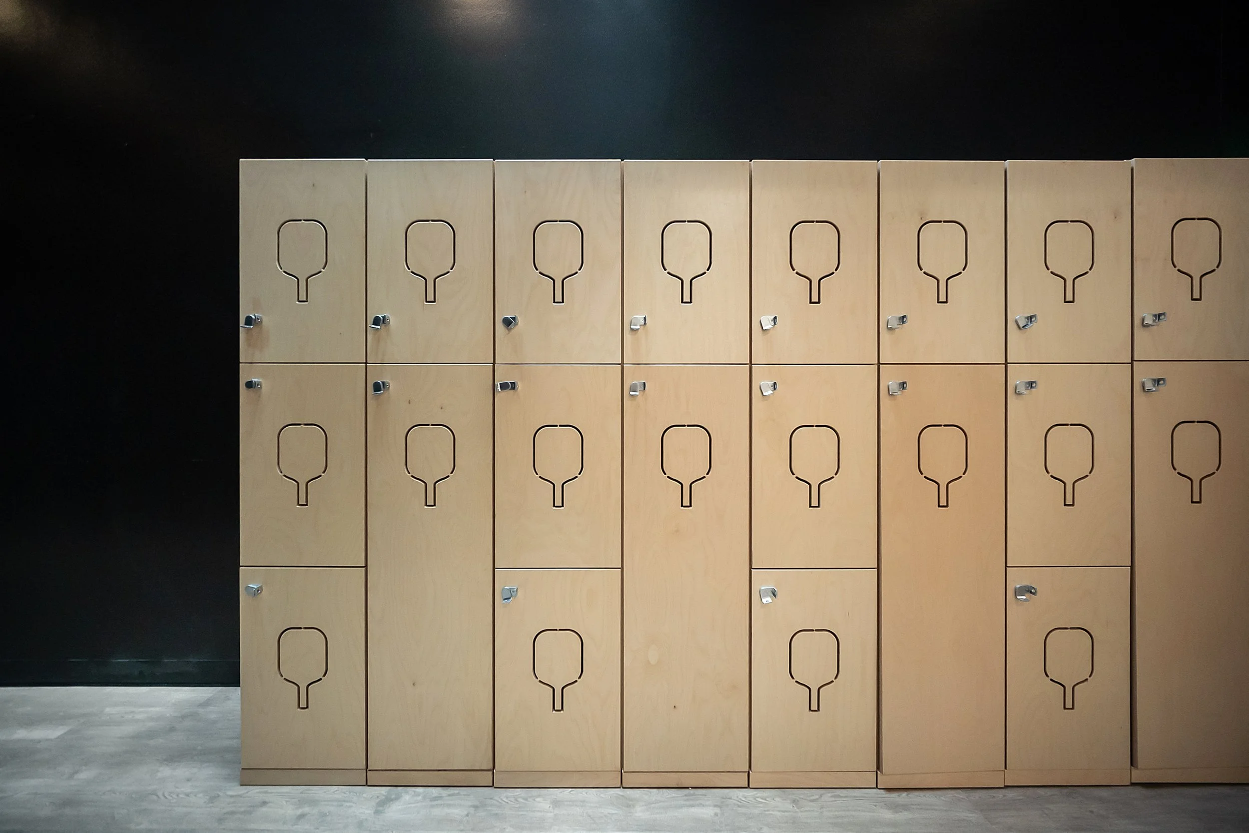 A large wooden locker unit with 16 compartments, each with a small lock, placed against a black wall with a light-colored floor.
