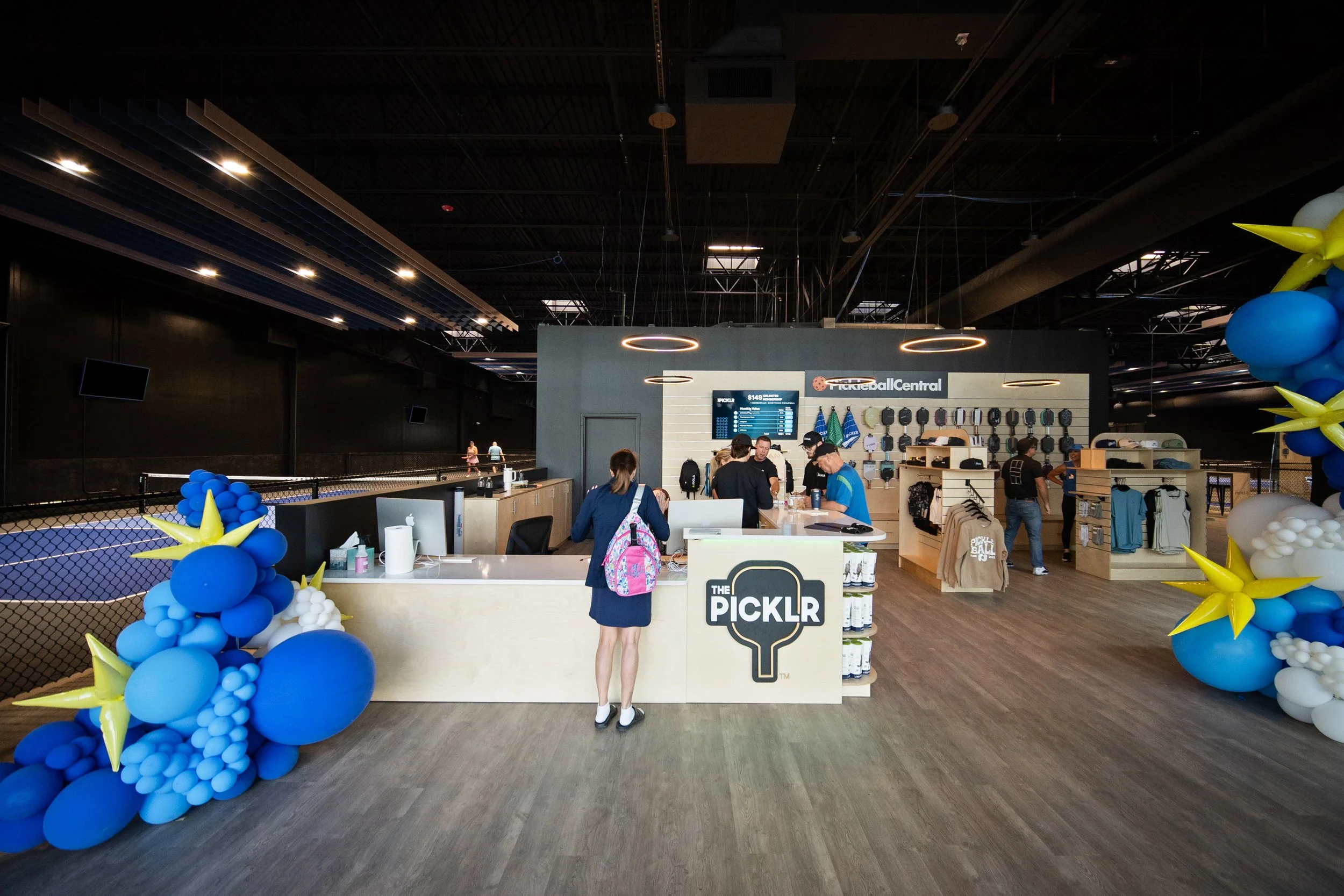 Inside a sports complex with a pickleball shop named The Picklr. There are blue and white balloon decorations, a counter with staff and customers, and a digital scoreboard displaying pickleball scores.