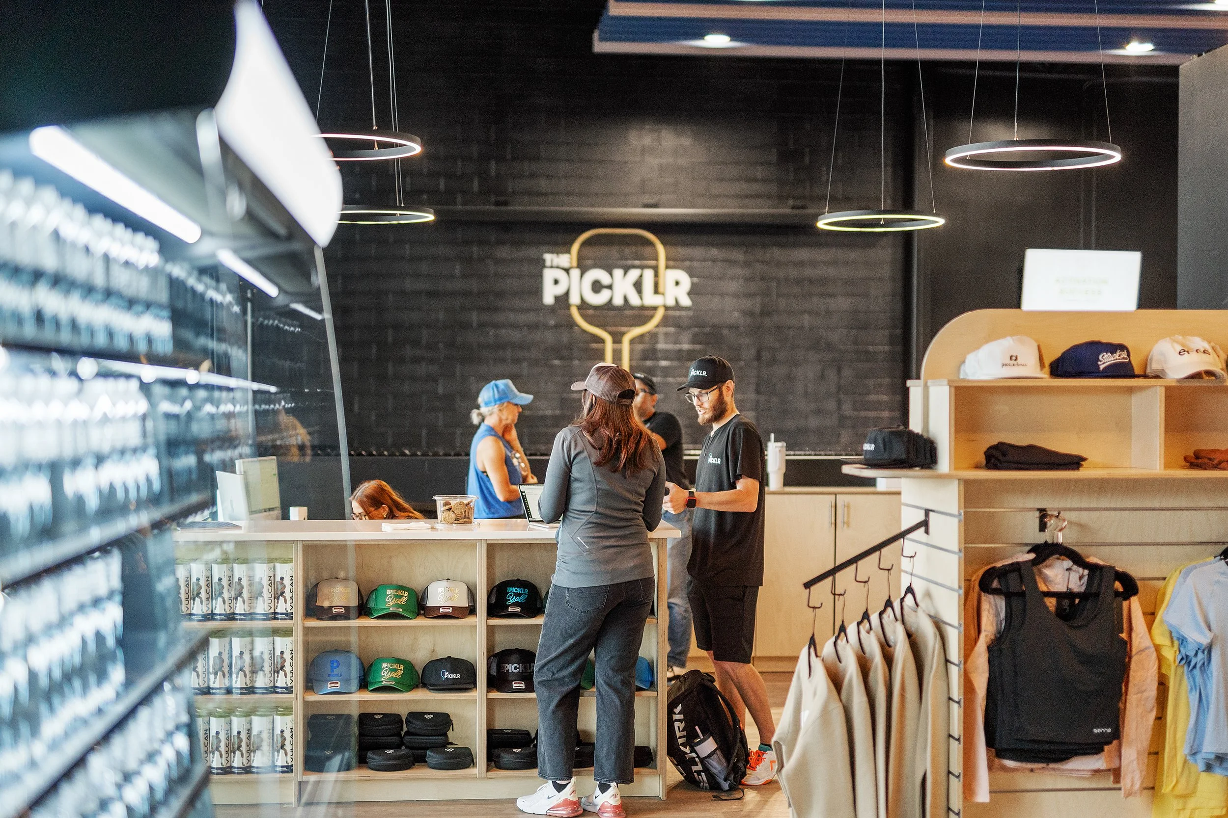 People at a store counter with a black brick wall background and a sign reading 'The PICKLR'. Shelves with hats and clothing are visible on the right side. Several customers and staff are engaged in conversation.