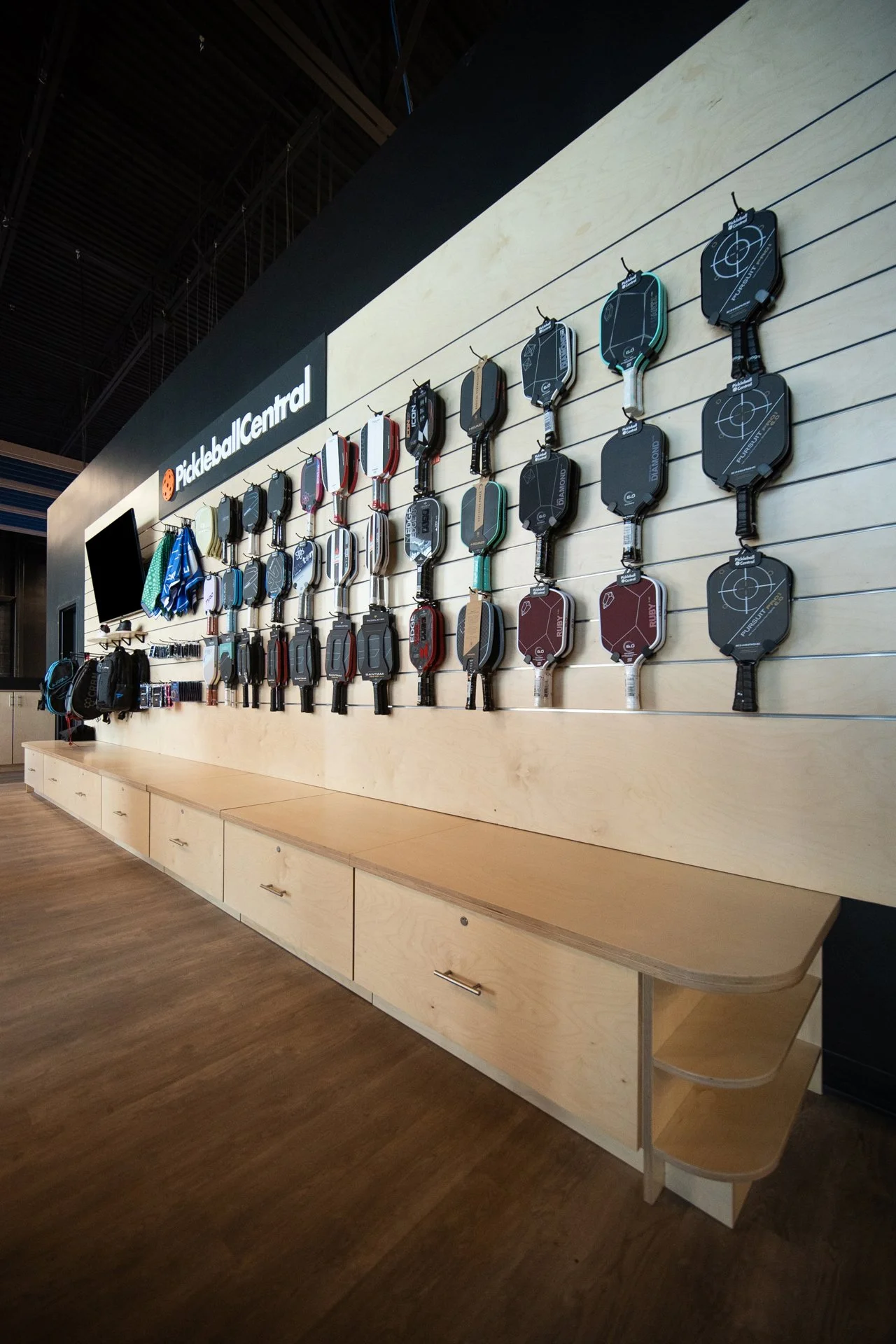 Display of pickleball paddles on a wall at Pickleball Central store, arranged in rows with different colors and designs, with a bench below for customers to sit or try out paddles.