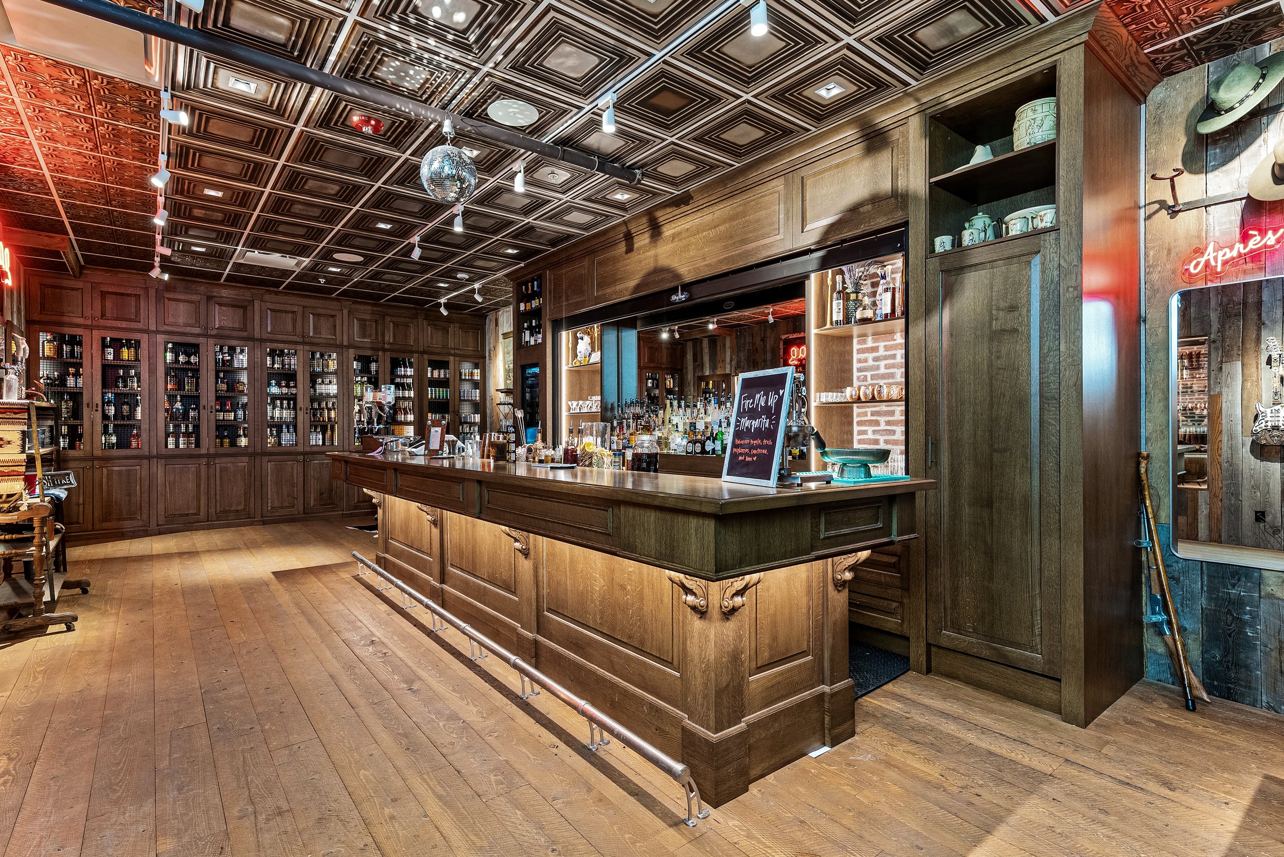 Interior of a rustic bar with a wooden counter, shelves of liquor bottles, a chalkboard menu, wood-paneled walls, and a decorated ceiling with spotlights and a disco ball.