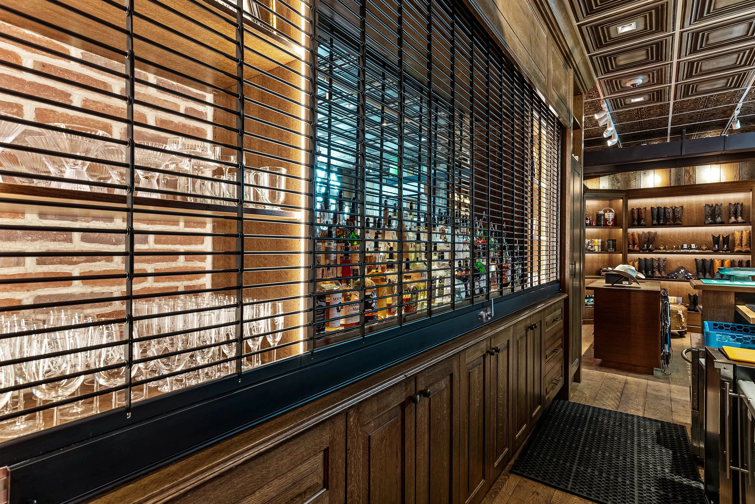 Interior of a bar or restaurant with a brick wall, wooden shelves with shoes or boots, and a glass display case filled with wine bottles. There is a black metal railing, a patterned ceiling, and warm lighting.