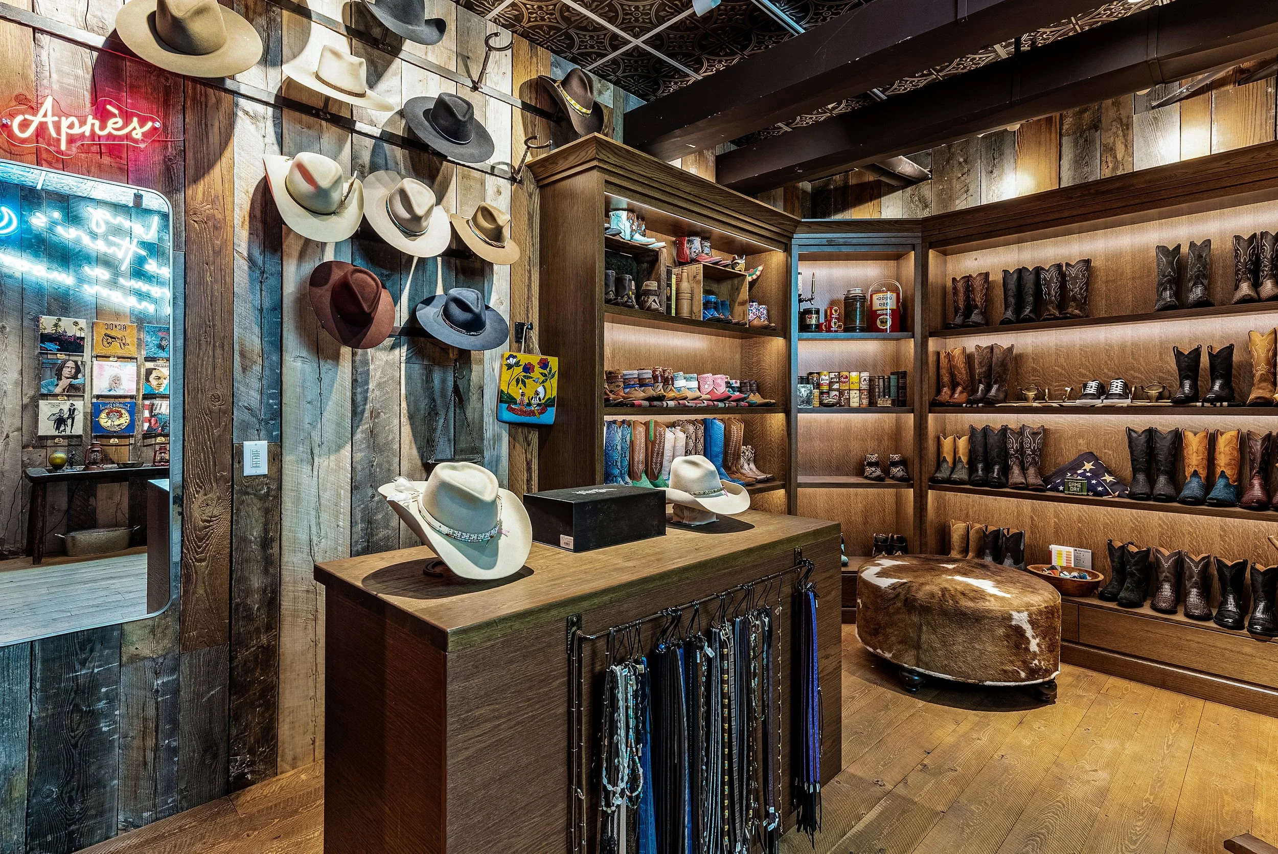 A store interior featuring a wall display of various hats, including fedoras and cowboy hats, and shelves with boots, jackets, and other accessories. There is a neon sign on the wall, and the overall decor has a rustic, Western theme with wood-panele