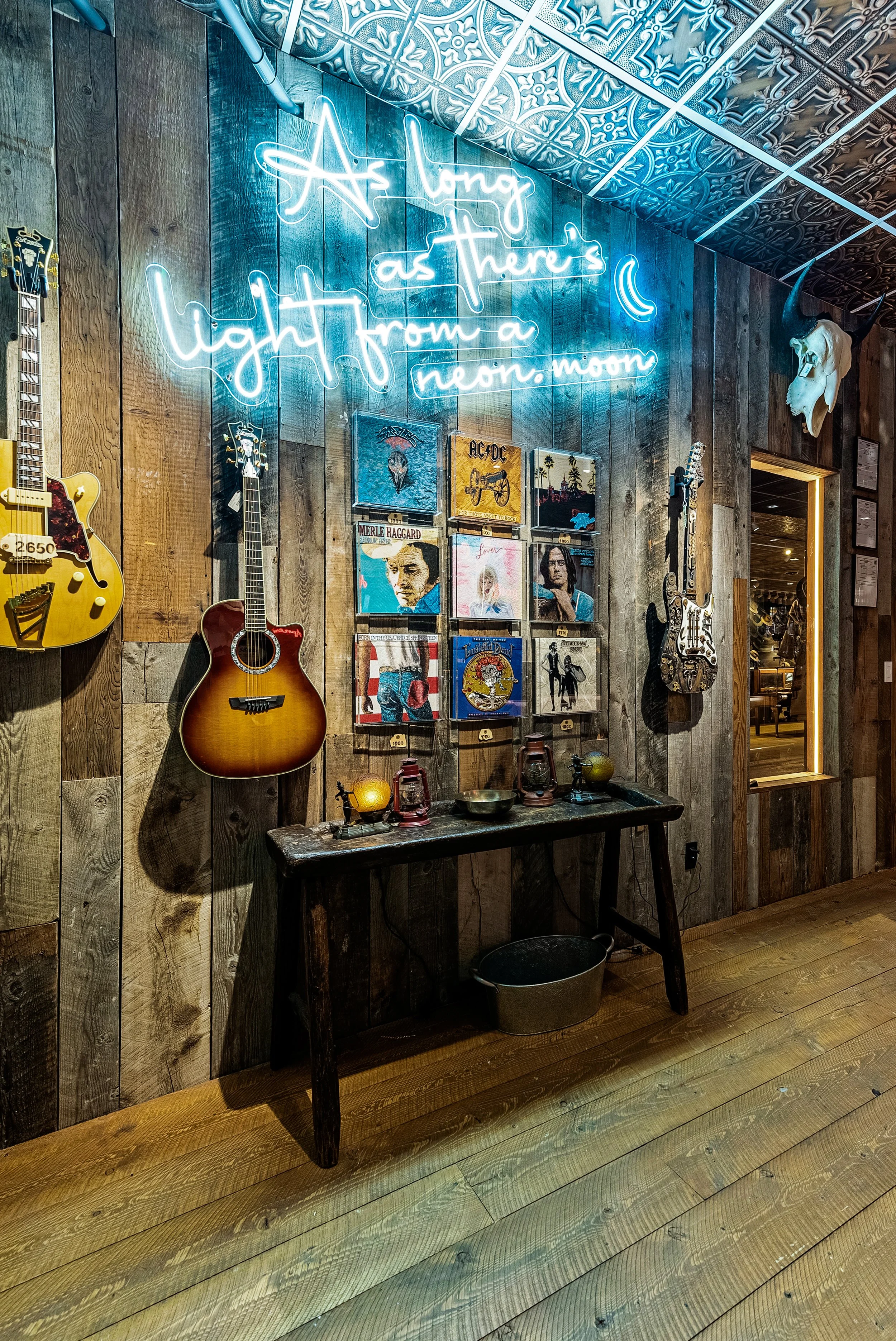 Interior of a rustic room with a wooden wall decorated with guitars, vinyl record album covers, and a neon sign that reads 'Among us light from a neon moon'. A small wooden table below the wall displays vintage lanterns and a potted plant. The ceilin