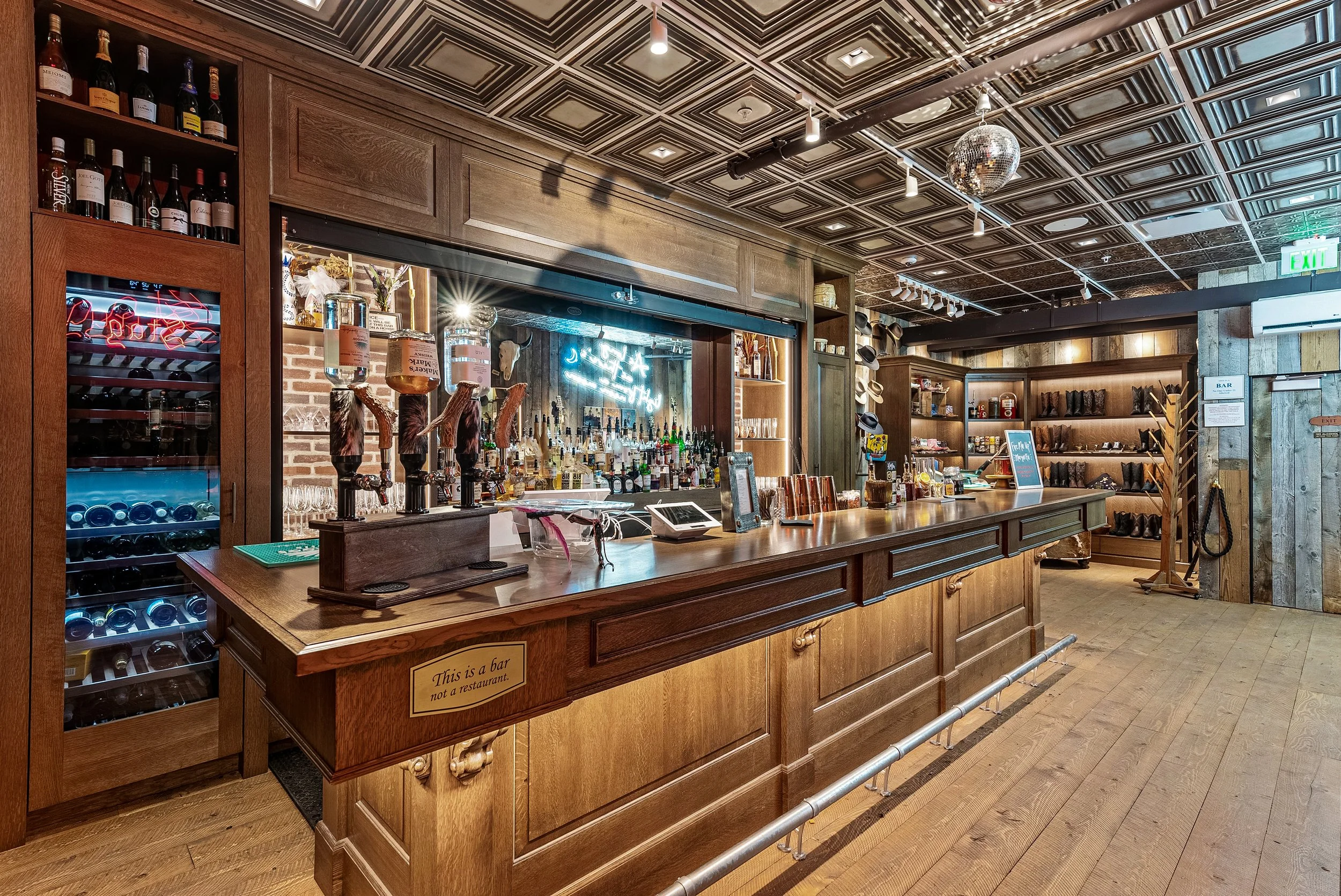 A bar inside a restaurant with a wooden counter, display of wine and liquor bottles, beer taps, and a neon sign. The ceiling has geometric designs and a disco ball. Shelves with hats and decorative items are in the background.