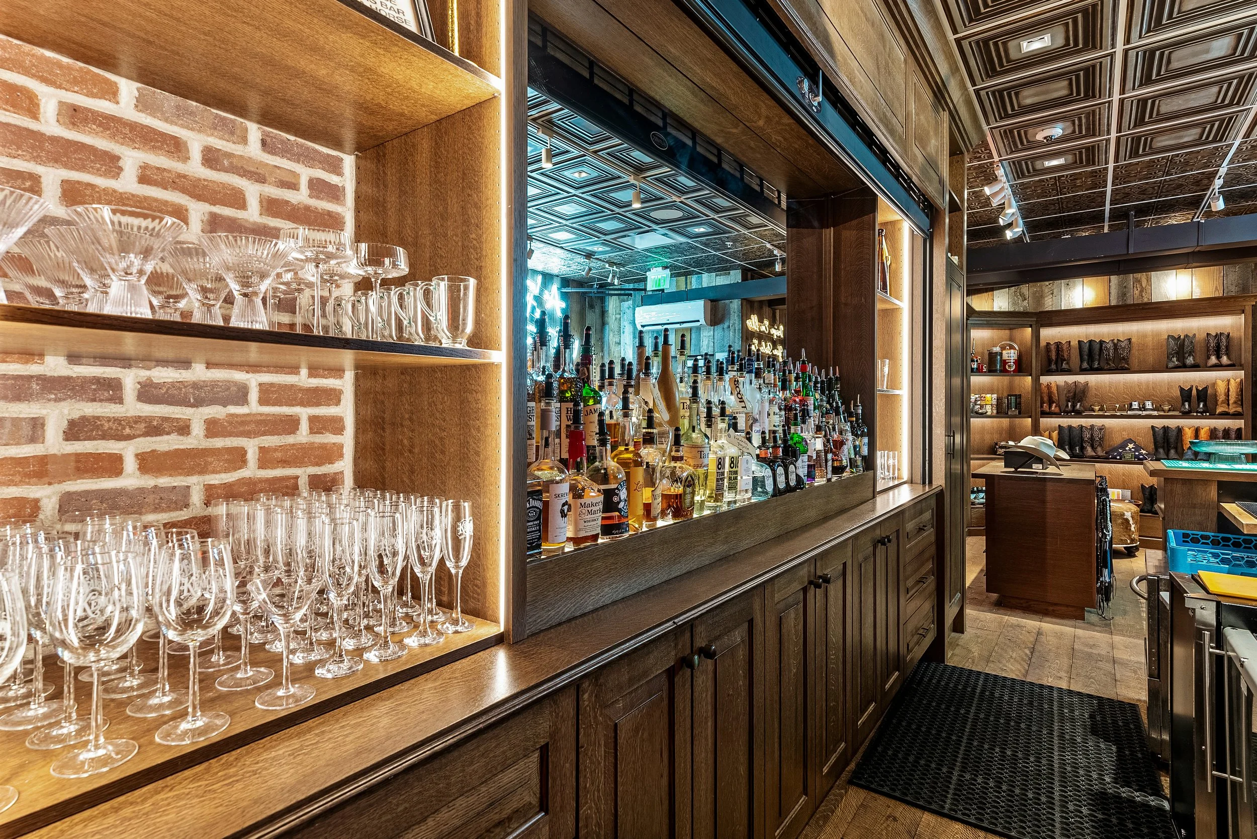 A bar area with shelves of glassware and liquor bottles, wooden cabinetry, and a decorated ceiling with track lighting in a cozy establishment.
