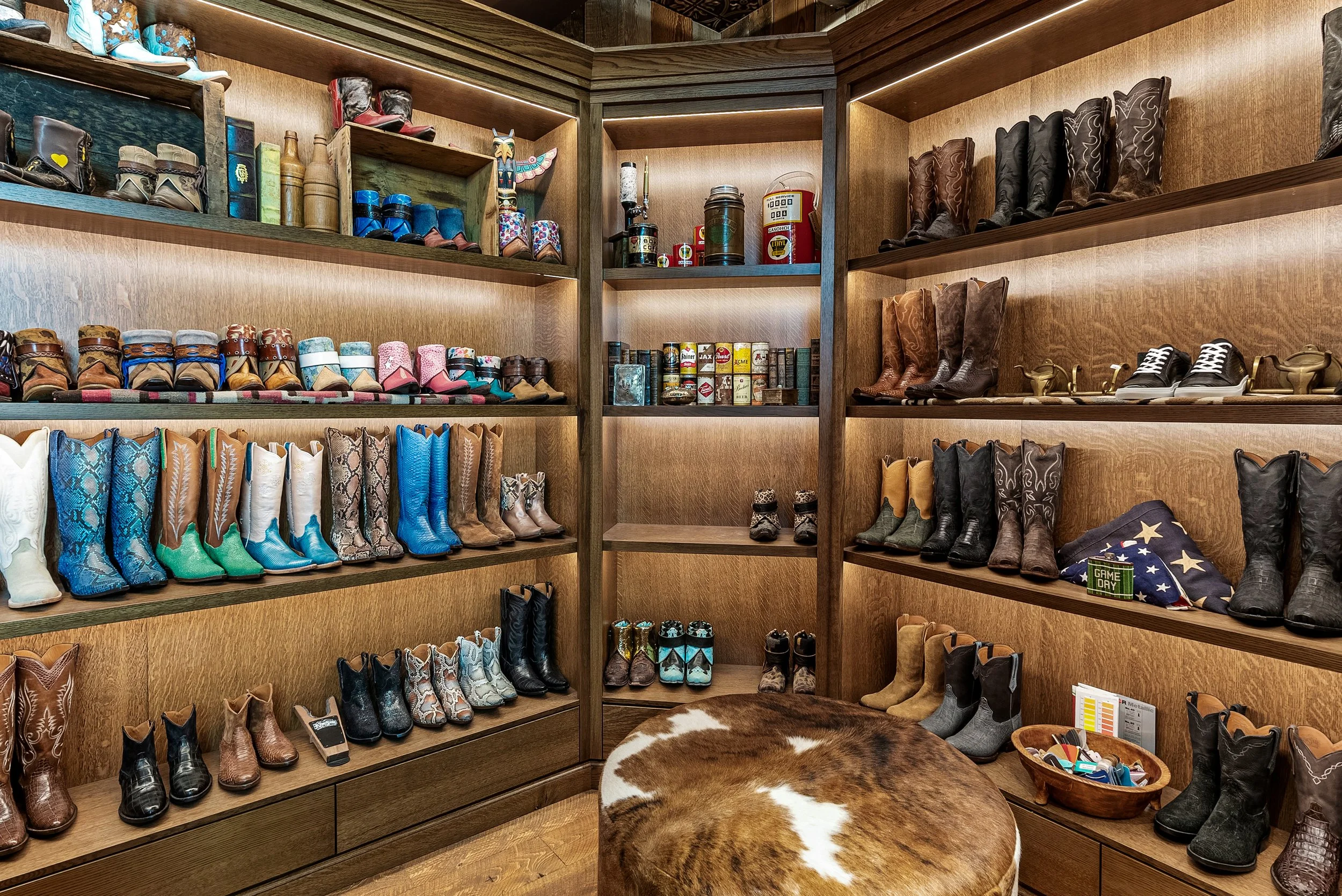 A closet with multiple wooden shelves displaying cowboy boots, shoes, and boots of various colors and styles. Some shelves also contain hats, books, storage tins, and decorative items.