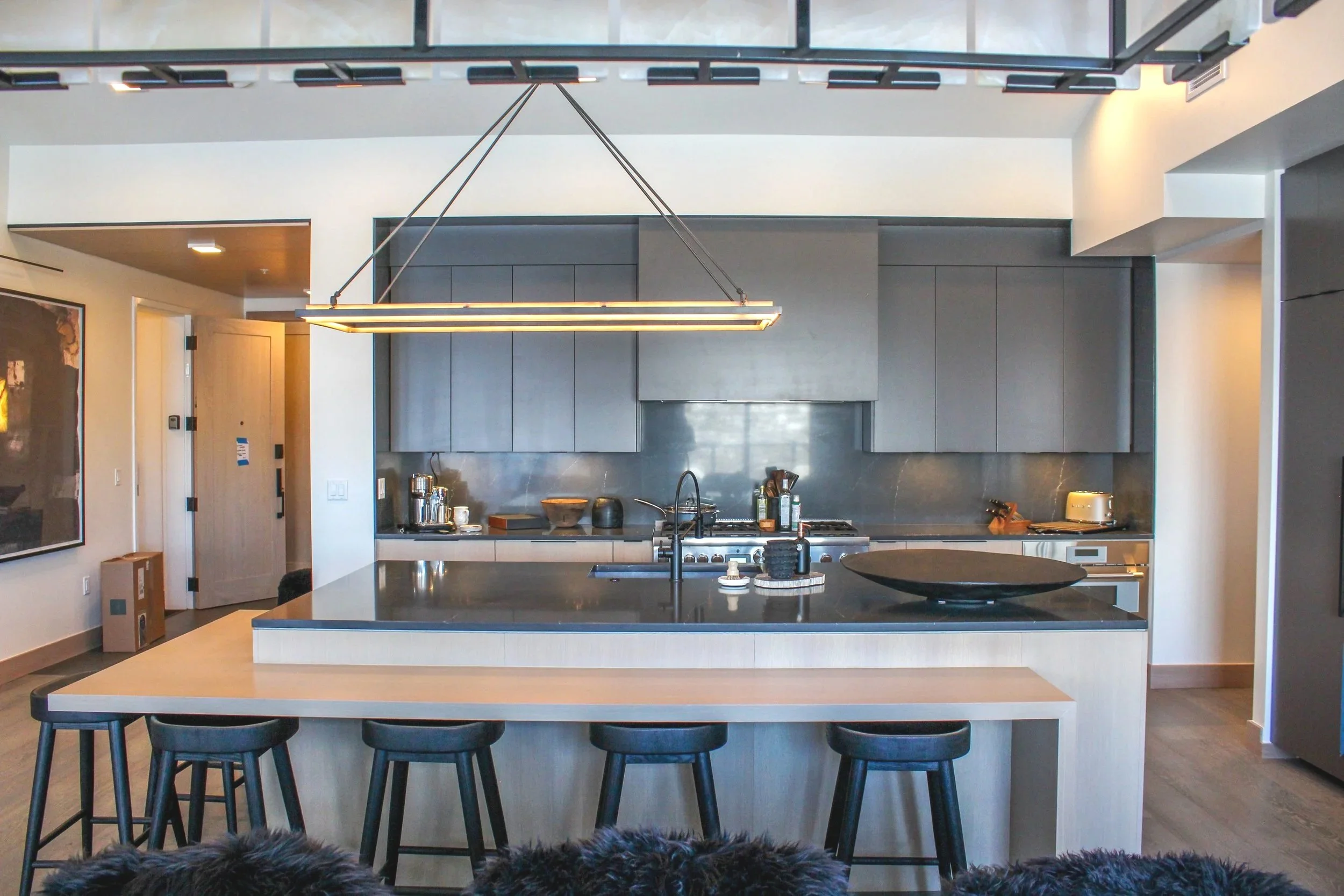 Modern kitchen with gray cabinets, black island, and chandelier, view from the living room with stools