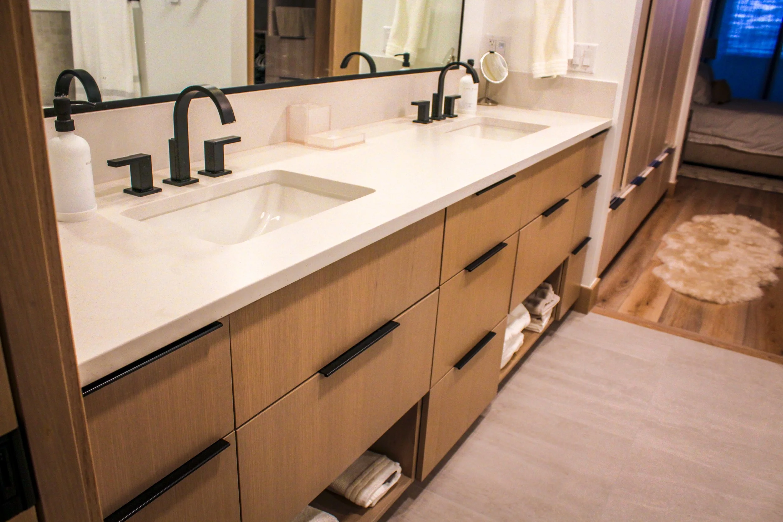 A bathroom vanity with a white countertop featuring three black sinks, each with a black faucet. A large mirror runs along the back of the vanity. To the right, there is a towel hanging on a hook and a mirror. Beneath the countertop, there are wooden