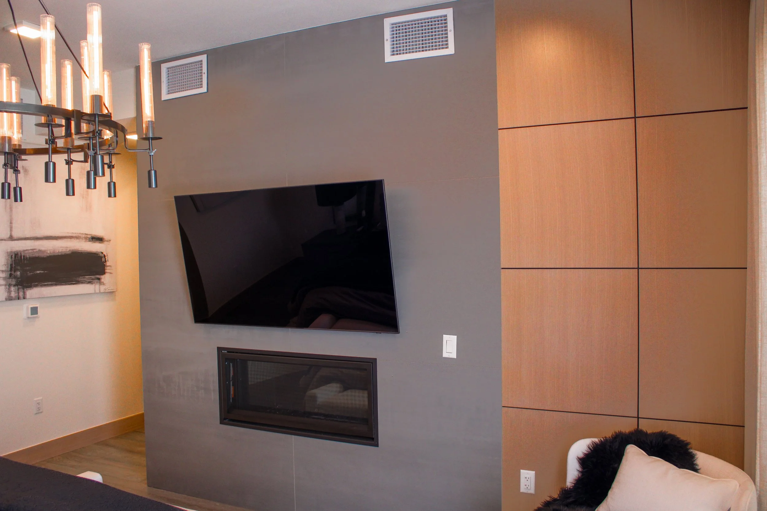 A modern living room corner with a wall-mounted flat-screen TV, a fireplace below it, and a beige chair with a black fur throw in front of a wooden paneled wall. A abstract painting hangs on an off-white wall to the left, and a contemporary chandelie